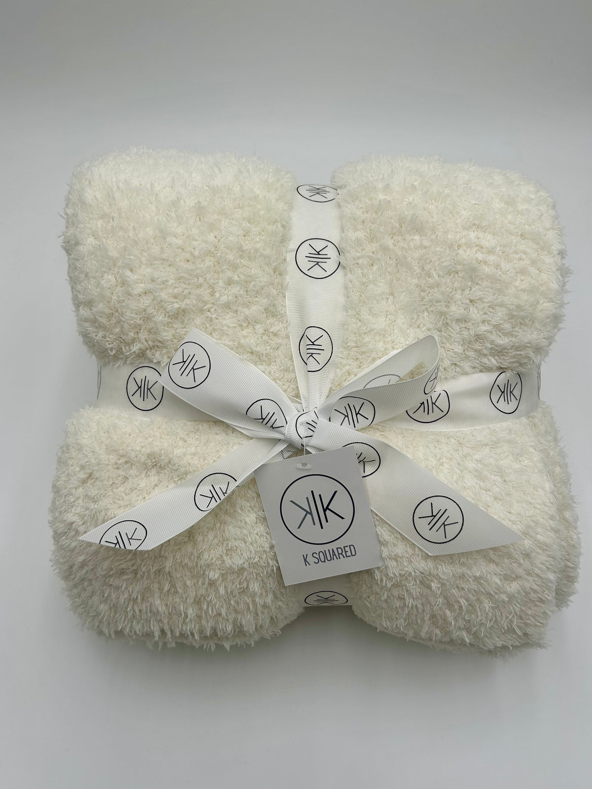 A folded cream-colored fluffy throw blanket tied with a white ribbon printed with the K Squared logo, on a light gray background.