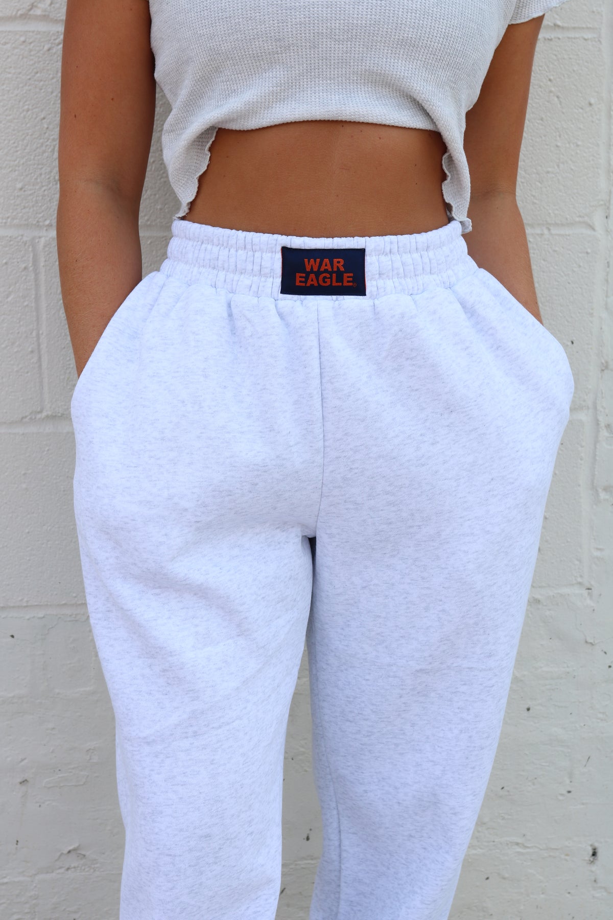 A person wearing light heather gray sweatpants with a visible War Eagle brand patch at the elastic waistband, paired with a cropped white top, standing against a white painted brick wall.