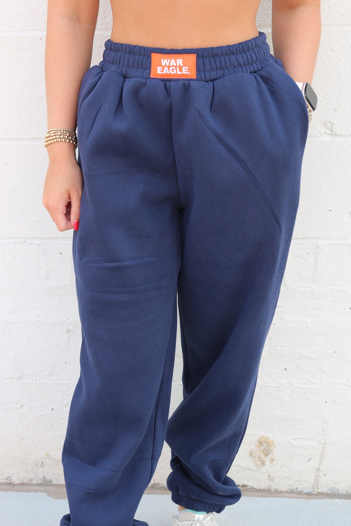 A person wearing navy blue sweatpants with an orange waistband patch that reads 'WAR EXCUSE' against a white painted brick wall.
