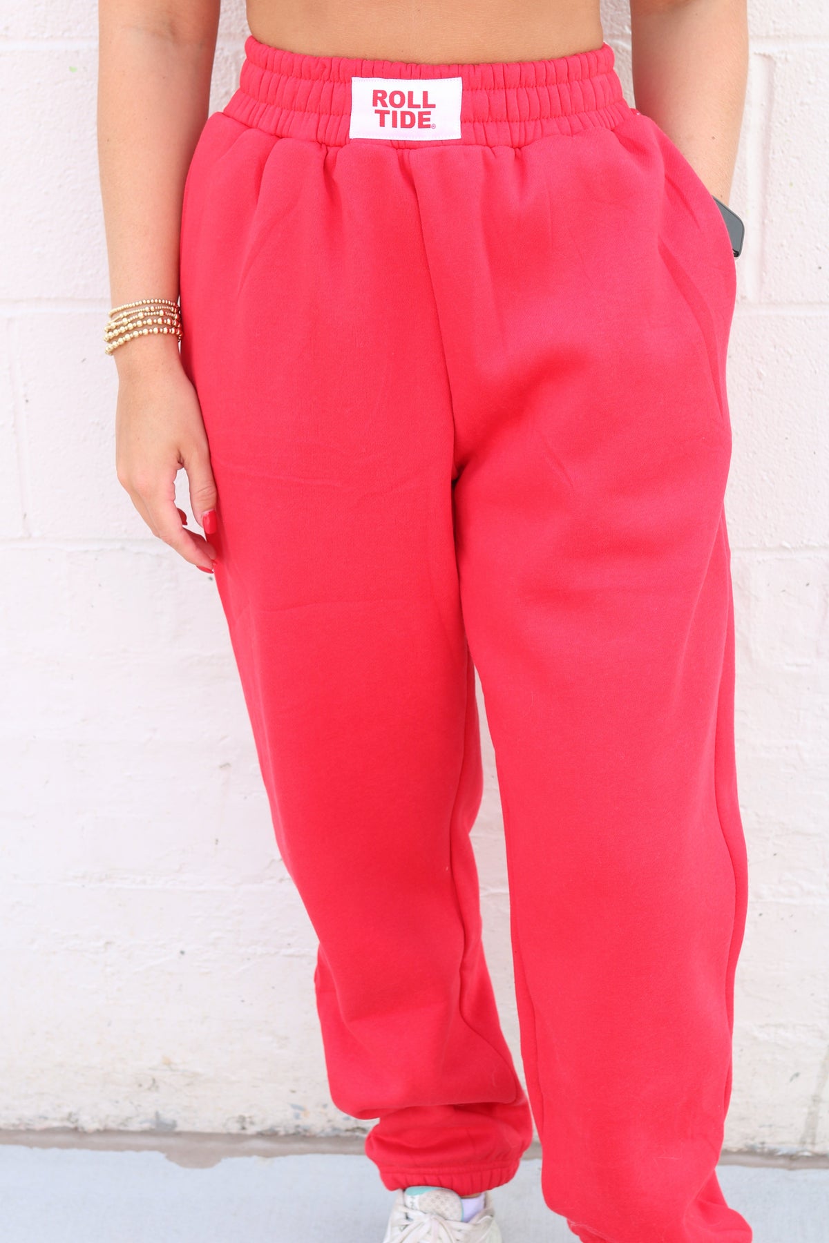 A person wearing bright coral pink sweatpants with an elastic waistband and a white 'ROLL TIDE' label patch, standing against a white brick wall.
