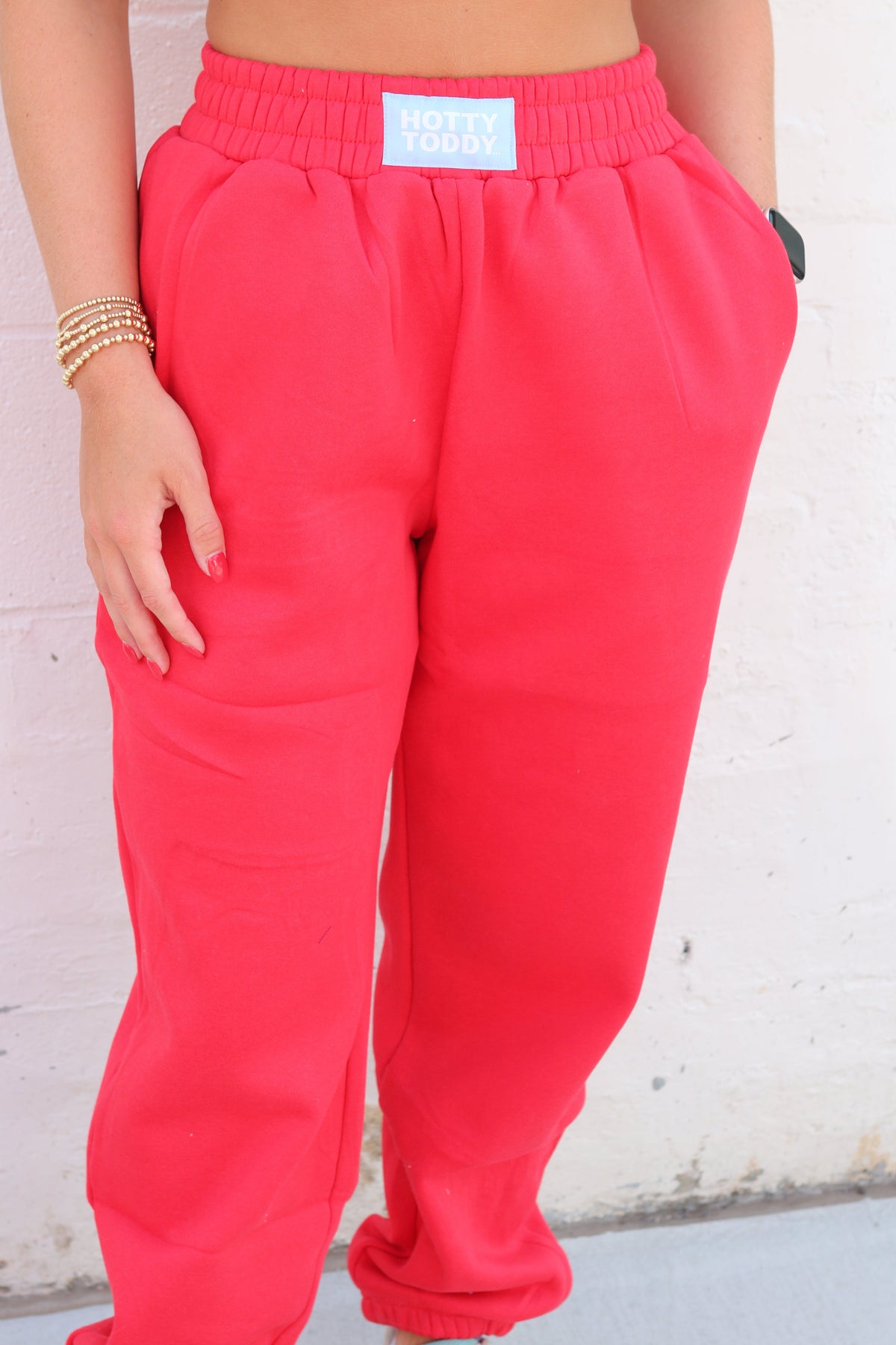 A person wearing bright coral-pink sweatpants with an elastic waistband and a small white brand patch reading 'NOTTY BRAND' at the center. The photo is cropped from the chest down, standing against a light painted brick wall.