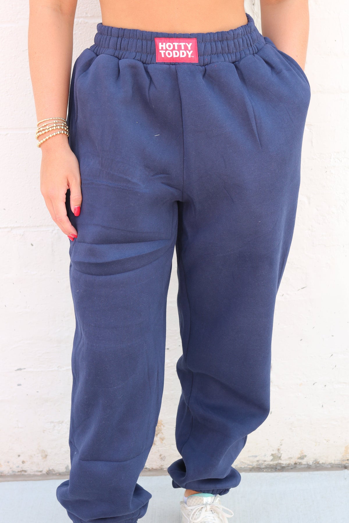 A person wearing navy blue sweatpants with an elastic waistband and cuffs, featuring a pink 'HOTTY TODDY' label at the back of the waistband. The person is also wearing white sneakers against a white brick wall background.