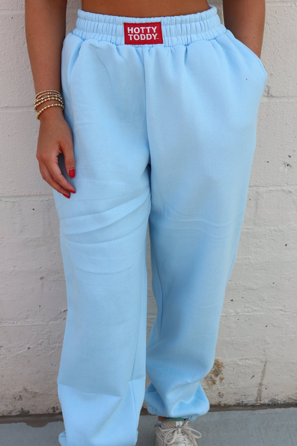 A person wearing light blue sweatpants with an elastic waistband and a red patch label reading 'HOTTY TODDY' on the front, standing against a white painted brick wall.
