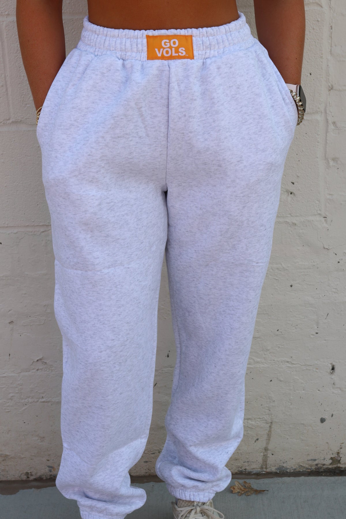 White sweatpants with an orange 'go vols' brand patch on the back waistband, worn by a model standing against a white brick wall.