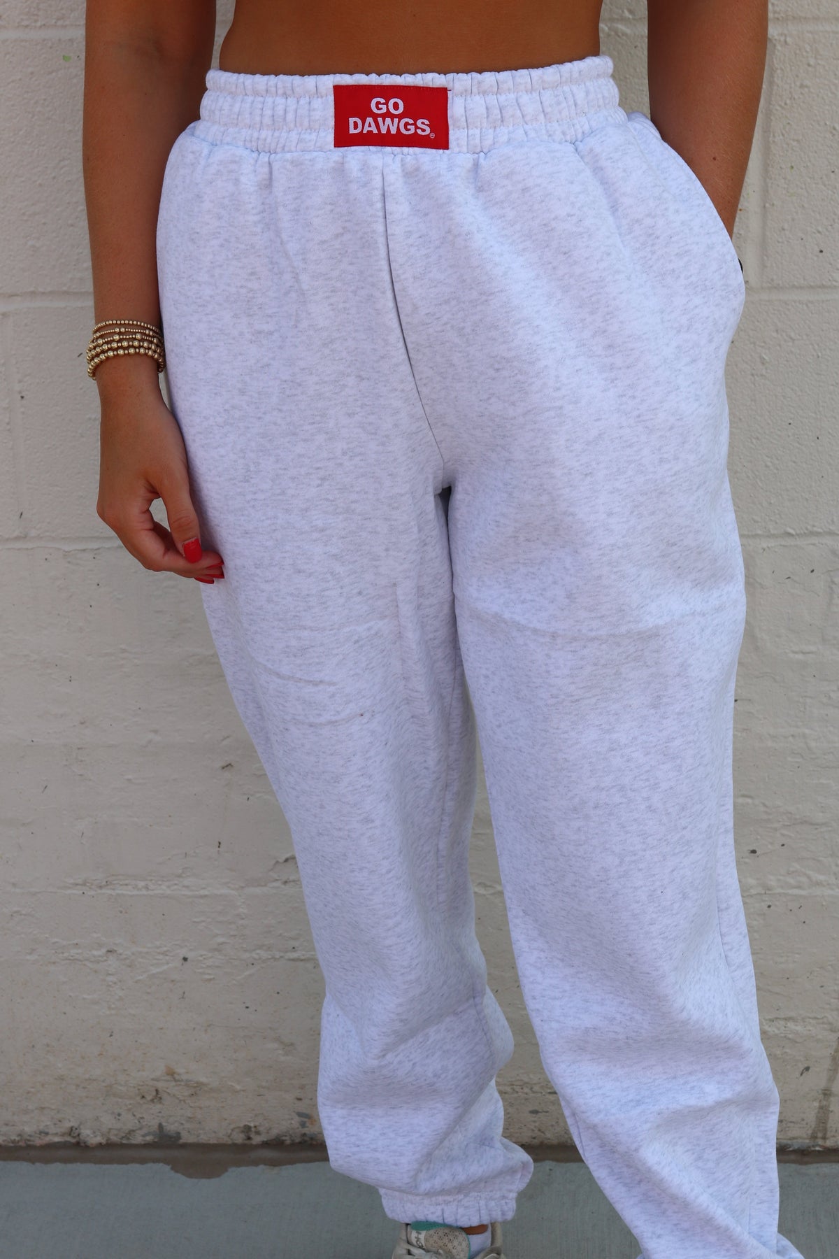 A person wearing light heather gray jogger sweatpants with an elastic waistband and a red 'GO DAWGS' patch on the front, standing against a white brick wall.