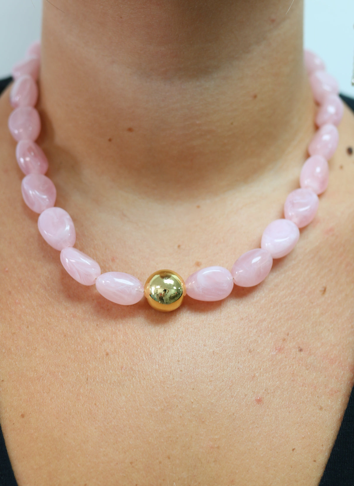A close-up of a person wearing a necklace made of pale pink polished beads with a single round gold bead at the center front.