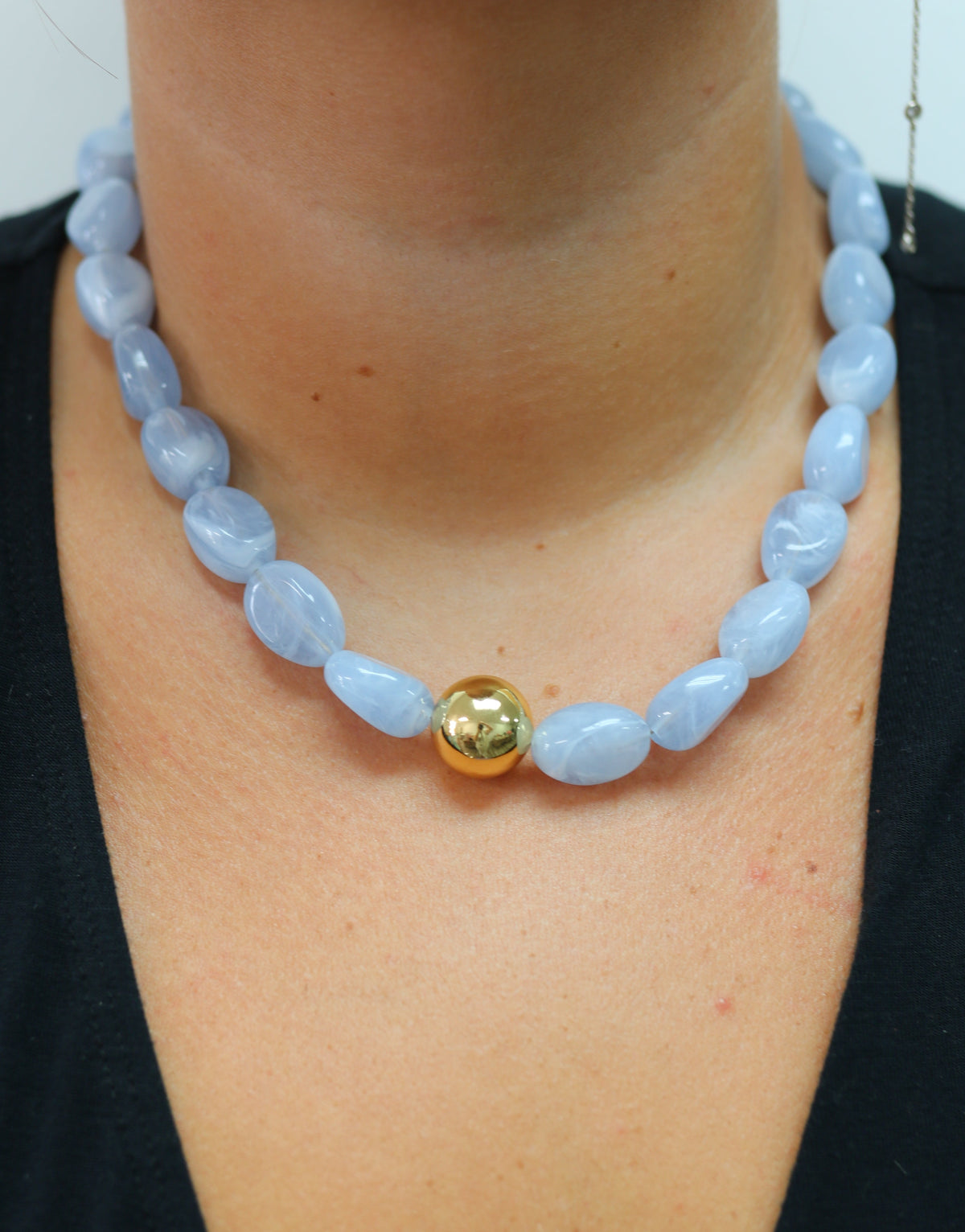 A close-up of a person wearing a light blue stone bead necklace with a single gold accent bead at the center, styled over a black top.