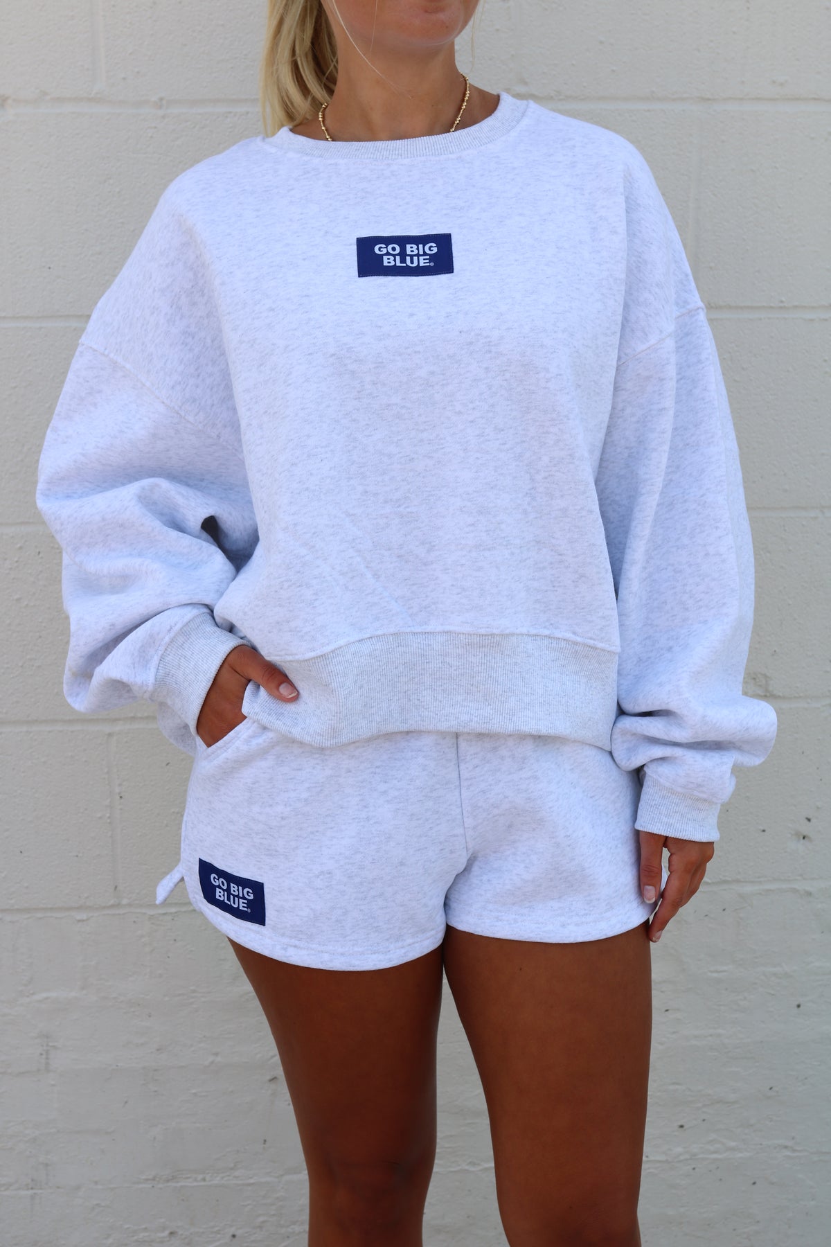 A person wearing a light heather gray sweatshirt and matching shorts set, photographed against a white painted brick wall. The outfit has small dark blue rectangular brand patches on the shorts and back of the sweatshirt.