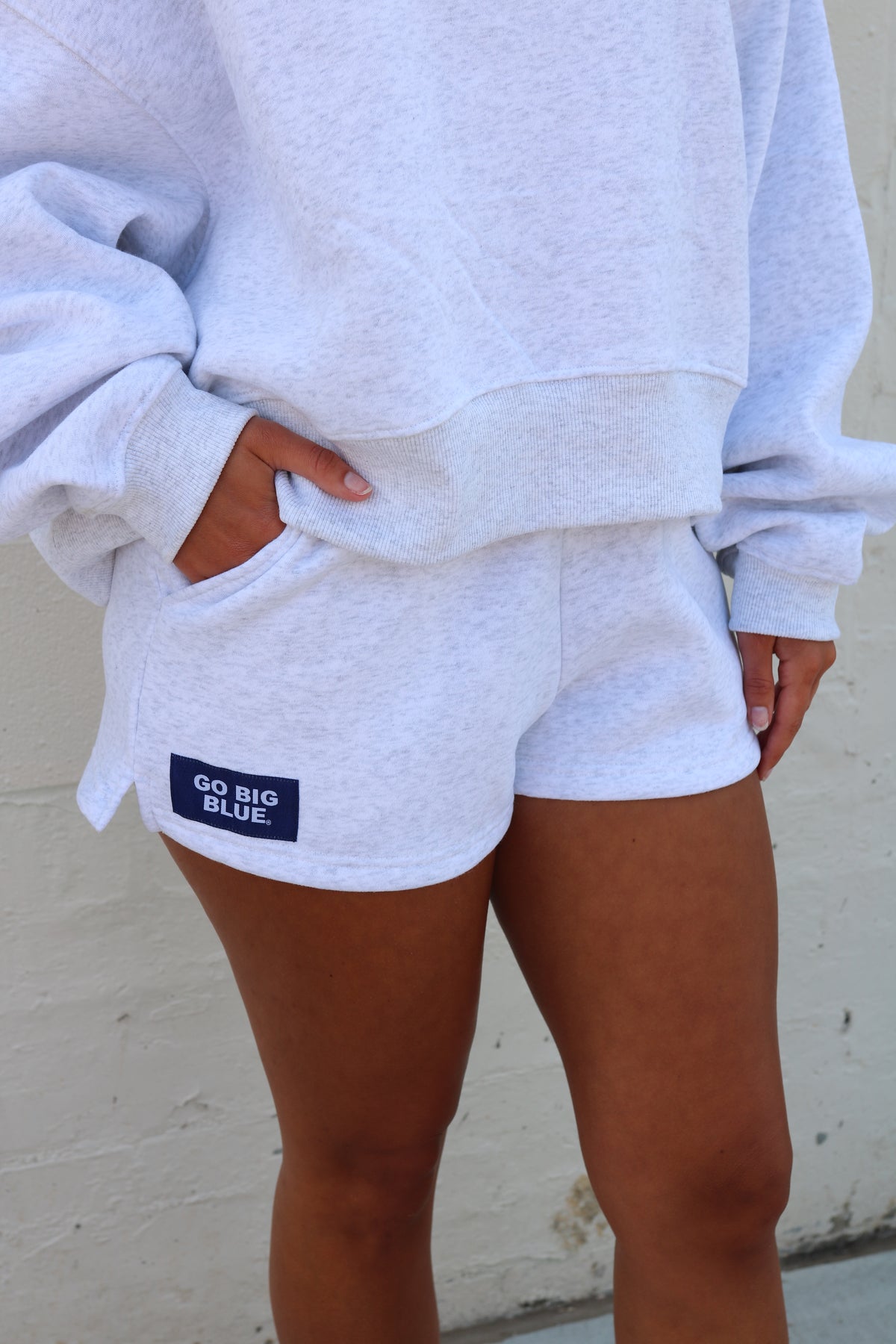 A person wearing light heather gray sweatpants with a side pocket and a small navy 'GO BIG BLUE' patch, paired with a matching light gray sweatshirt, standing against a light textured wall.