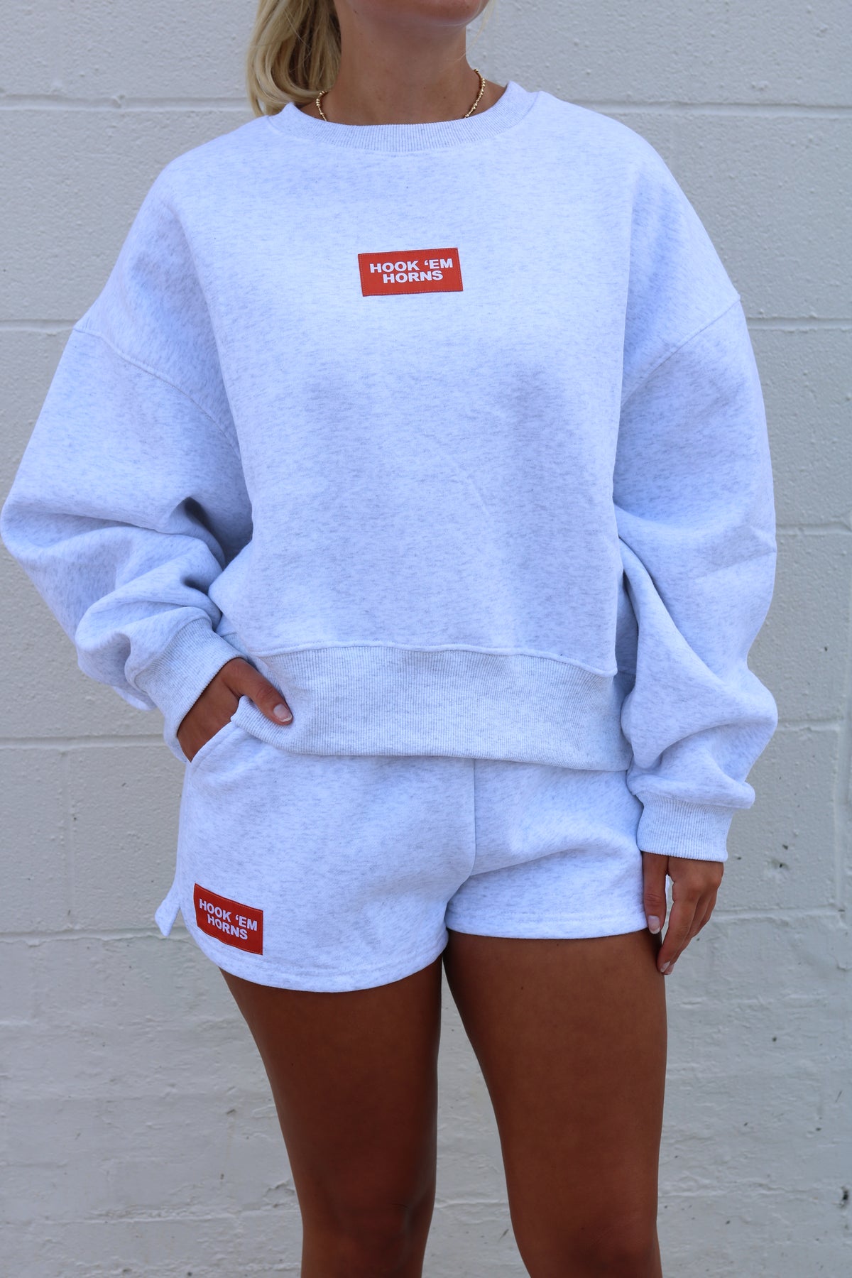 A woman wearing a matching light heather gray sweatshirt and shorts set with red rectangular chest and thigh patches that read 'HOOK 'EM MOMMY'.