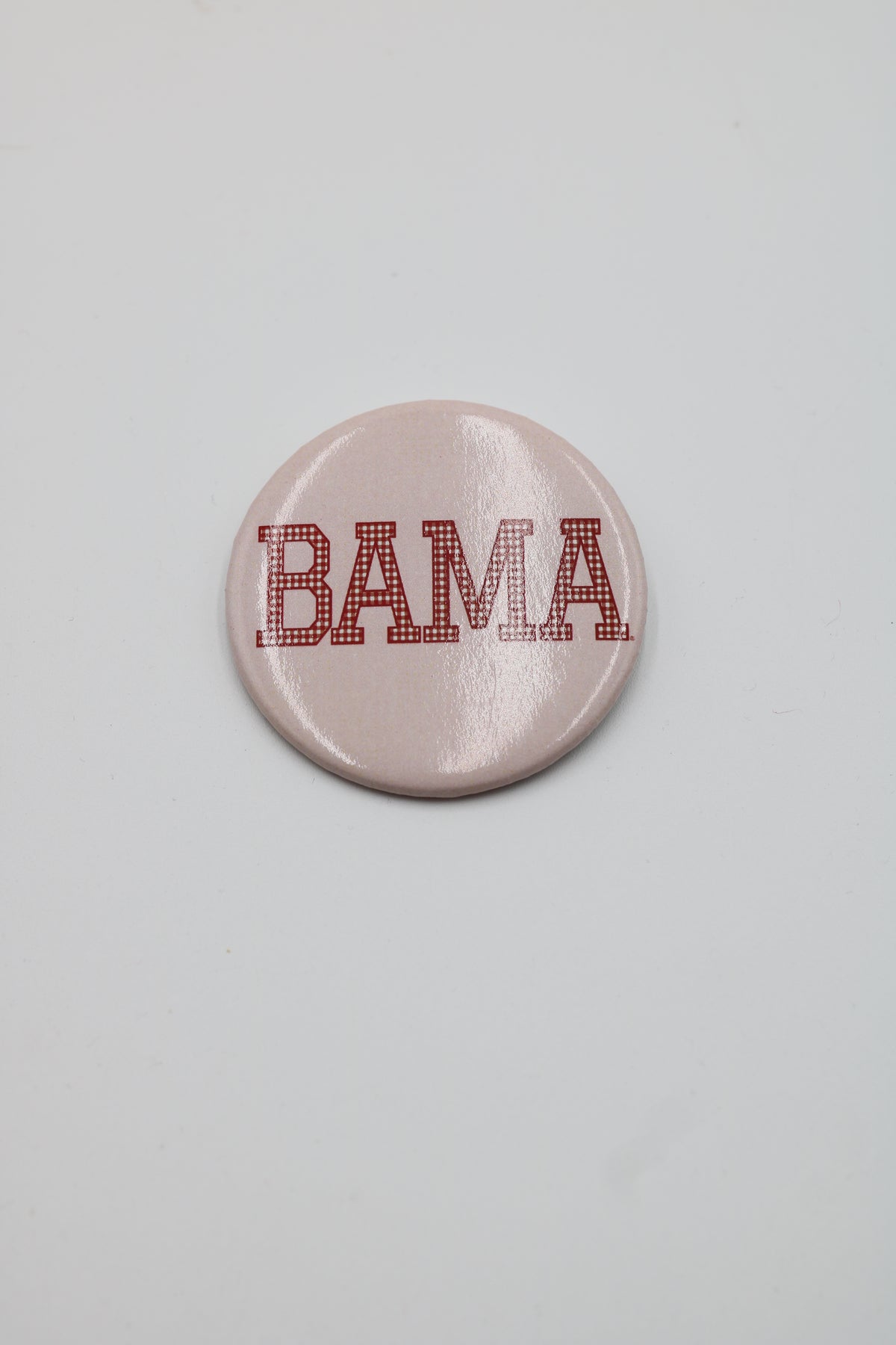A round pin-back button badge with a glossy light pink background and the word 'BAMA' in red block letters outlined with small white dots, centered on a plain light gray background.