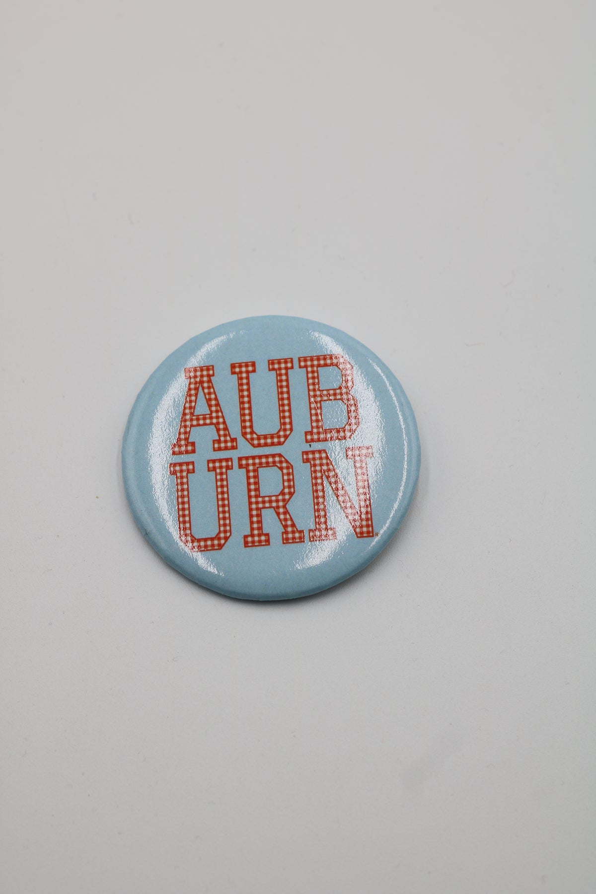 A round pinback button badge with a light blue background and red text reading 'AUBURN' in a dotted font, shown on a plain light gray background.