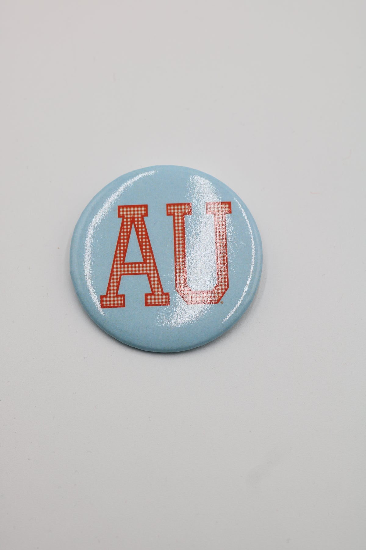 A round light blue button pin featuring the red-orange letters 'AU' in a gingham pattern, set against a plain light gray background.