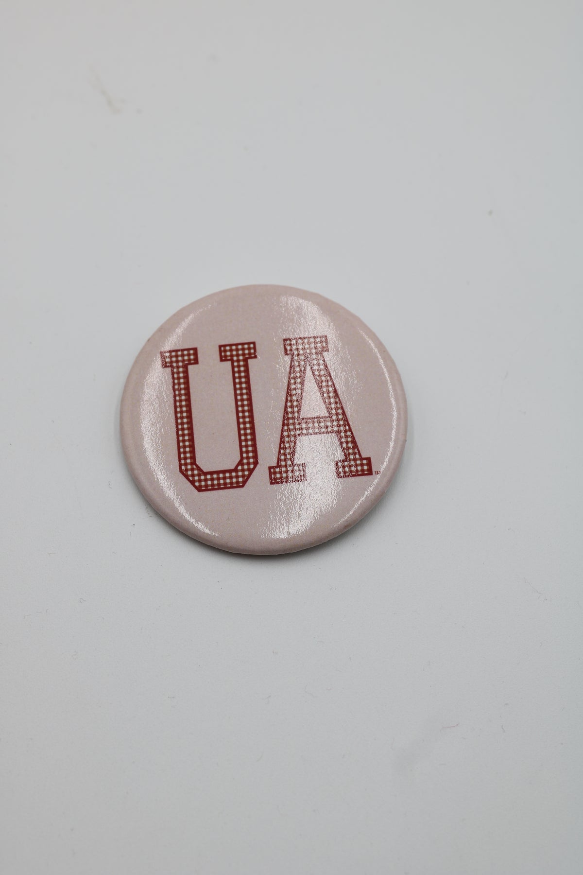 A round pinback button badge featuring the letters 'UA' in a red dotted collegiate font on a light pink background, photographed on a plain light gray surface.