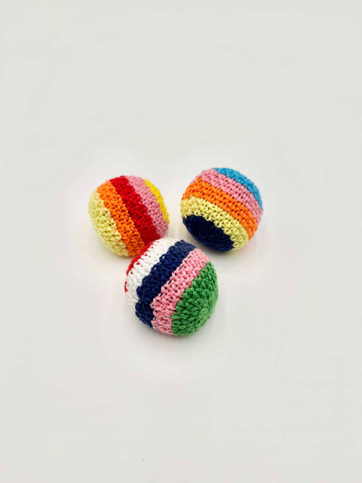 Three small crocheted striped balls in rainbow colors, arranged on a plain light gray background.