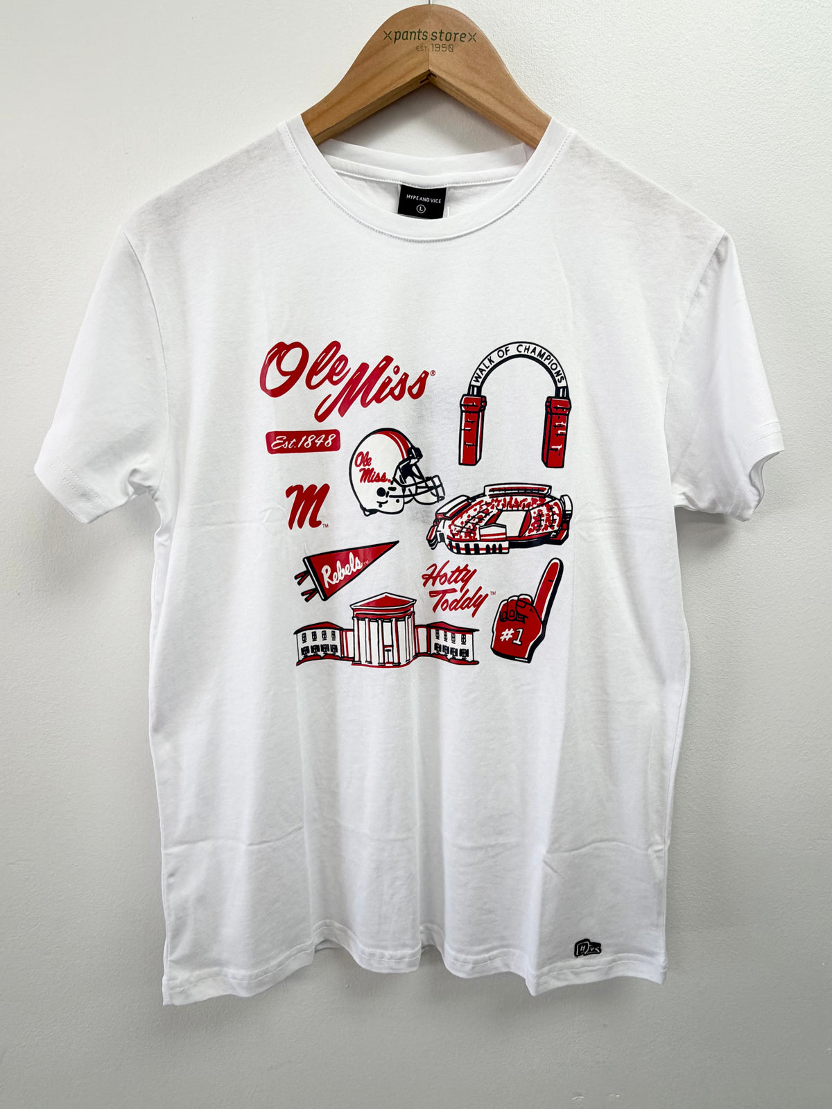 A white short-sleeve t-shirt hanging on a wooden hanger against a light gray wall. The shirt features a red and black graphic print on the front with the words 'Ole Miss', 'Belle Star', 'M', 'Hasty Reddy', a football helmet, a car, and other collegiate-themed illustrations.