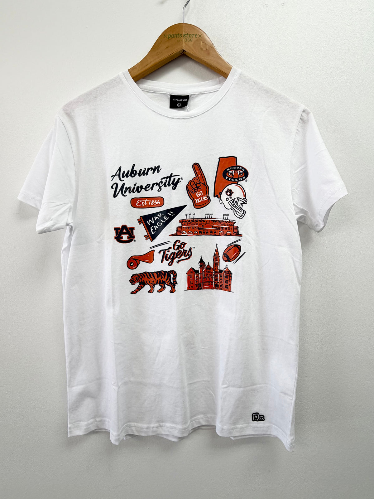 A white short-sleeve t-shirt hanging on a wooden hanger against a light gray wall. The shirt features a graphic print on the front with Auburn University branding, including the text 'Auburn University CHICAGO', a football helmet, a football, a tiger, and a pennant in red, orange, and black.