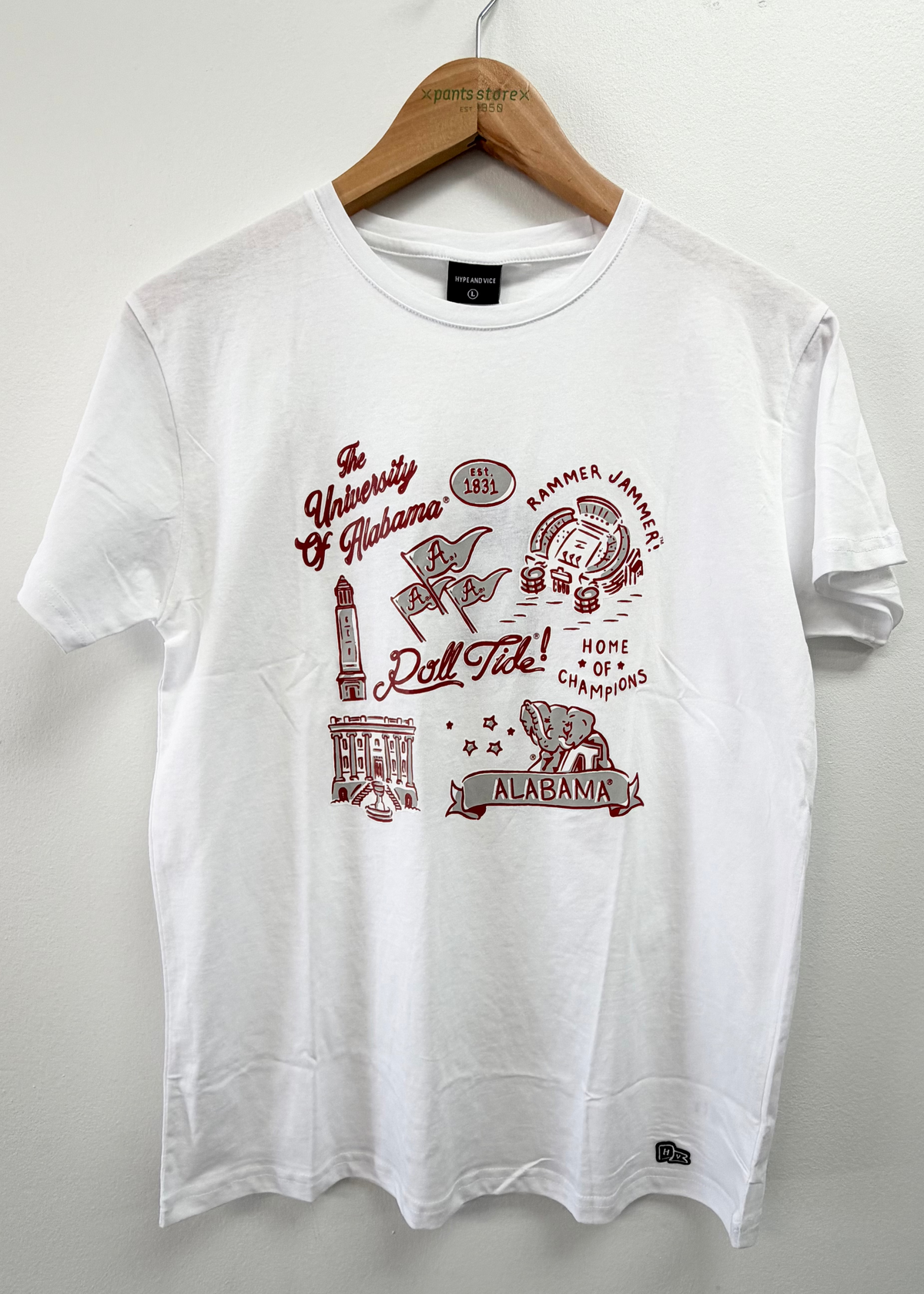 A white short-sleeve t-shirt hanging on a wooden hanger against a light gray wall. The shirt features a maroon graphic print on the front with collegiate and sports-themed illustrations and text, including 'The University of Alabama', 'Alabama', and 'Roll Ste.