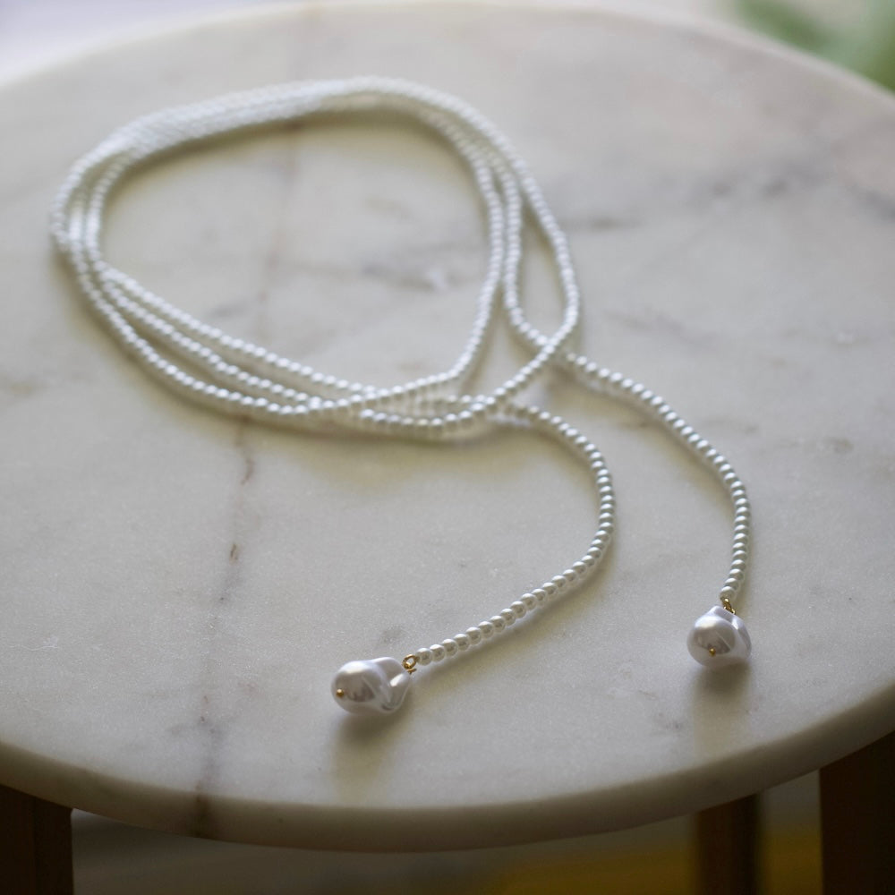 A delicate silver beaded necklace with two large teardrop-shaped silver beads at the ends, displayed on a light marble surface.