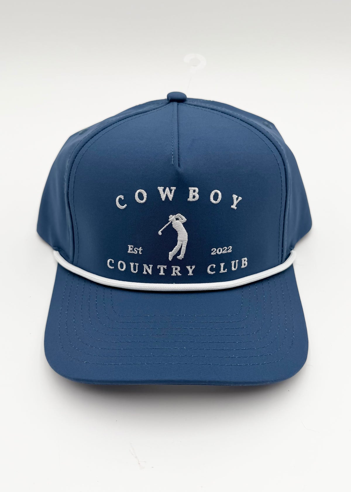 A blue baseball cap with white embroidered text reading 'COWBOY COUNTRY CLUB' and a small golfer graphic on the front, plus a white piping detail around the brim, shown on a light gray background.