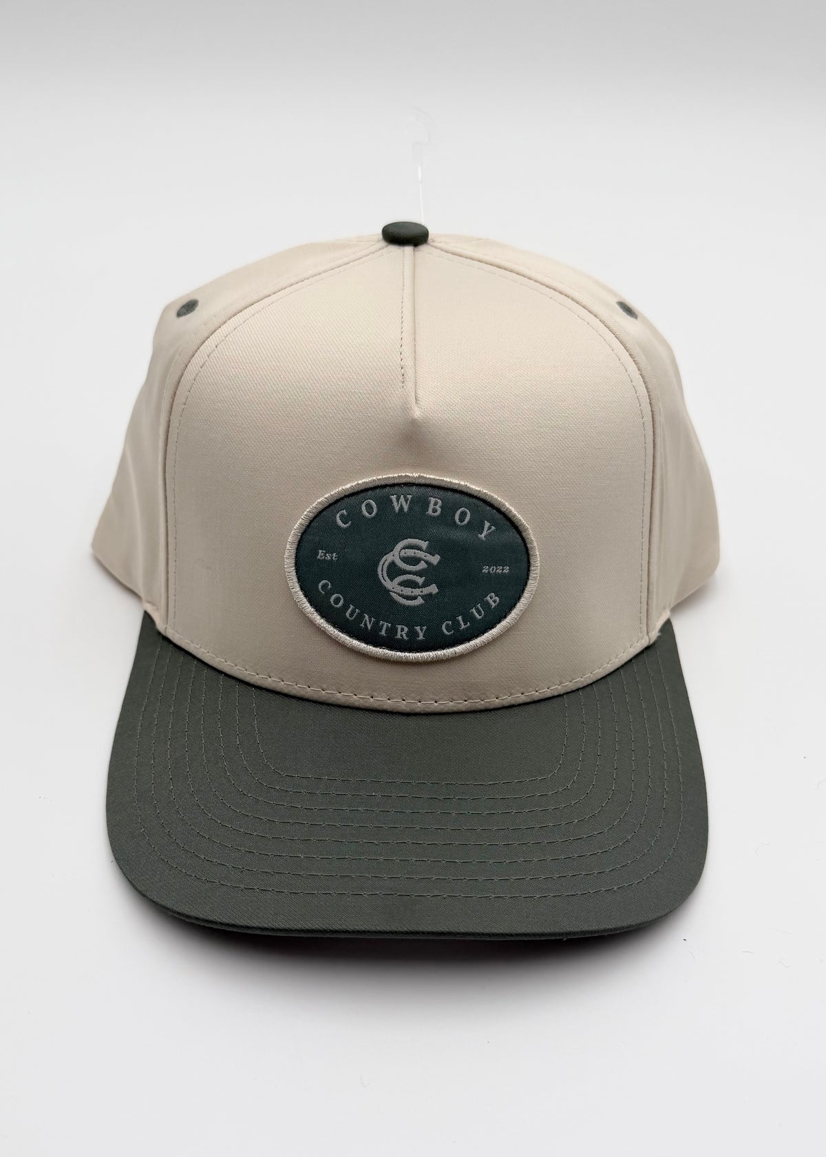 A structured baseball cap with a light beige crown and a dark gray flat brim, featuring an oval patch on the front that reads 'COWBOY COUNTRY CLUB'.