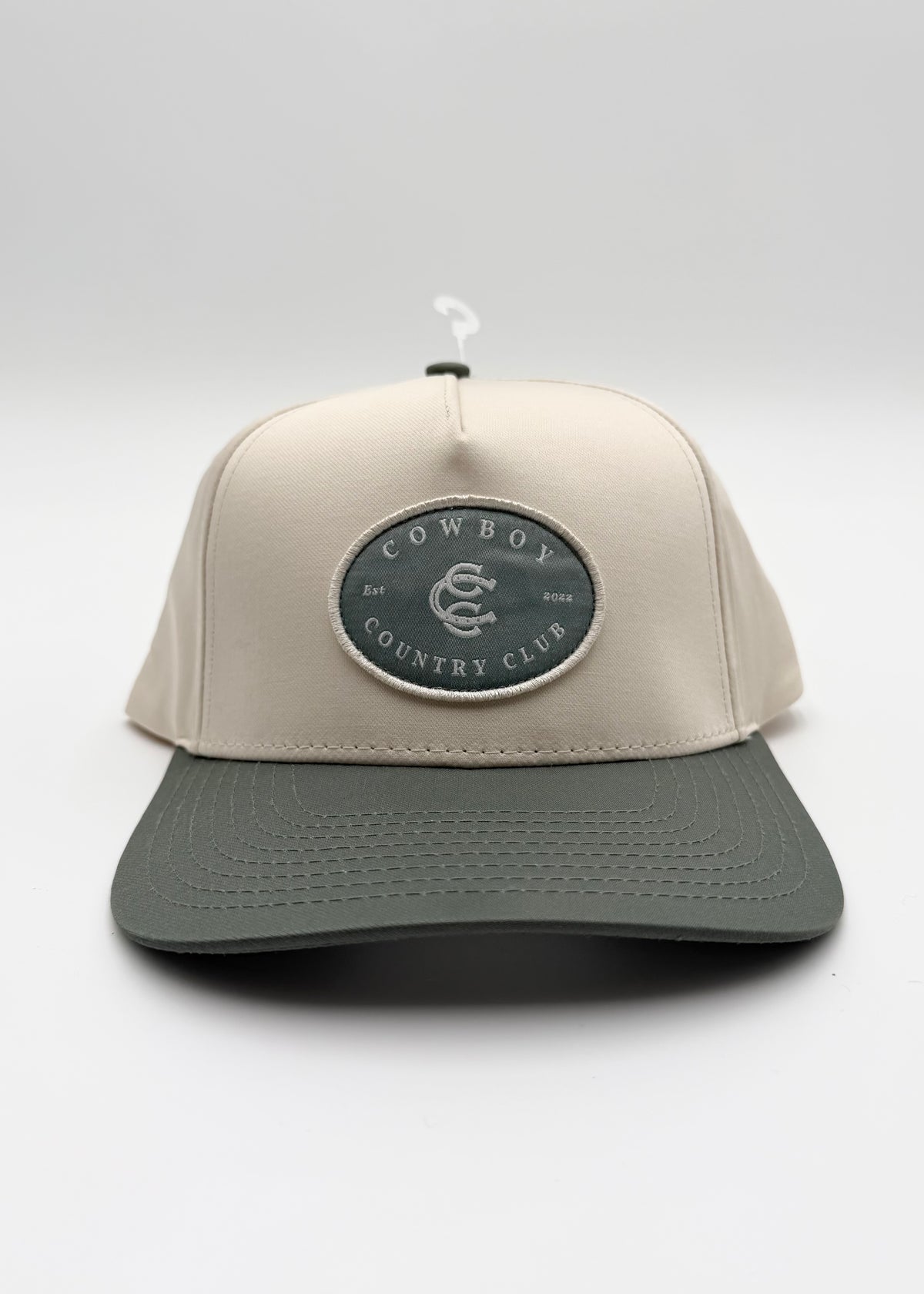 A structured baseball cap with a light beige crown and a dark gray brim, featuring a circular embroidered patch on the front that reads 'COWBOY COUNTRY CLUB'.