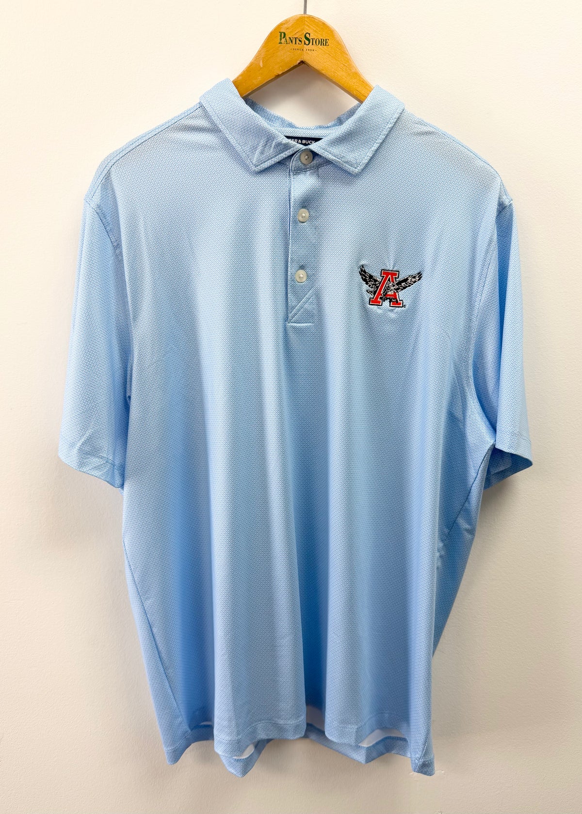A light blue short-sleeve polo shirt hanging on a wooden hanger against a plain beige wall. The shirt features a small embroidered logo on the left chest with a red and black eagle design.