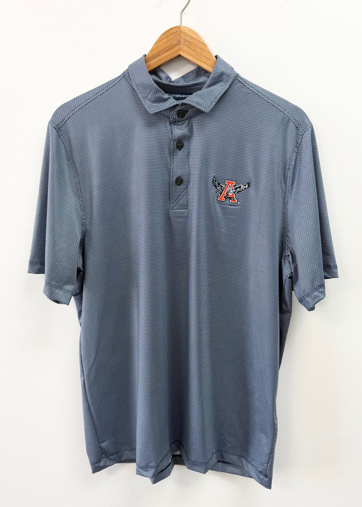 A slate blue short-sleeve polo shirt with a small embroidered logo on the left chest, hanging on a wooden hanger against a plain white wall.