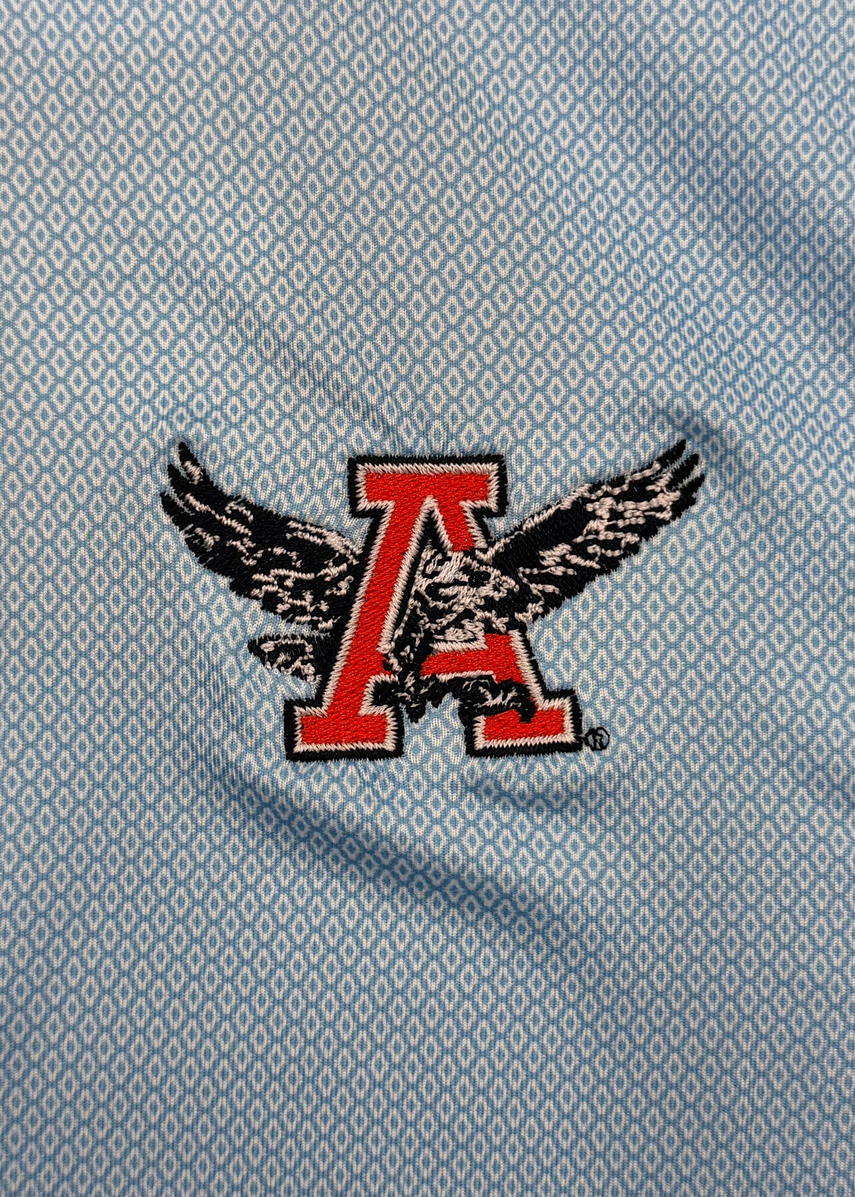 A close-up of a light blue patterned fabric with a red and black embroidered 'A' logo featuring an eagle design.