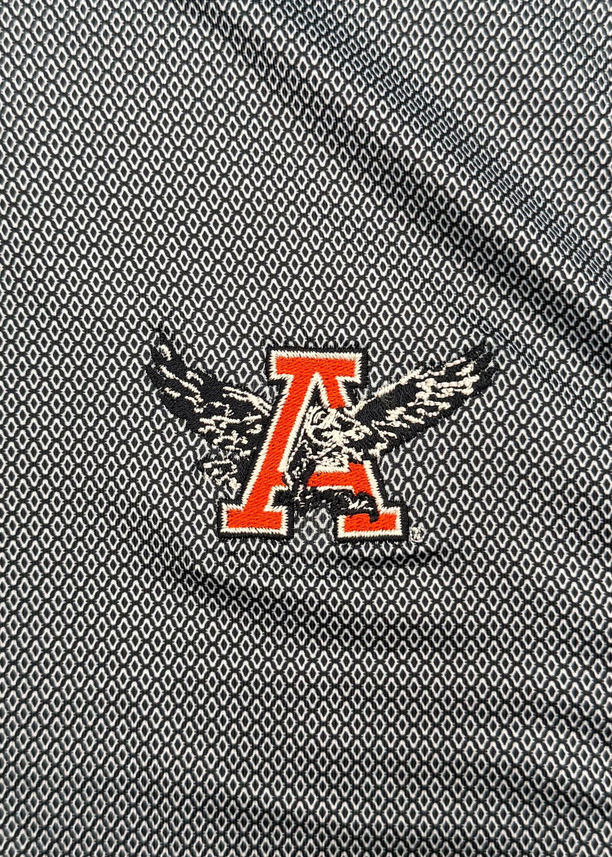 A close-up of a textured gray fabric featuring a repeating geometric pattern and a central graphic of a red letter 'A' overlaid with a black and white eagle design.