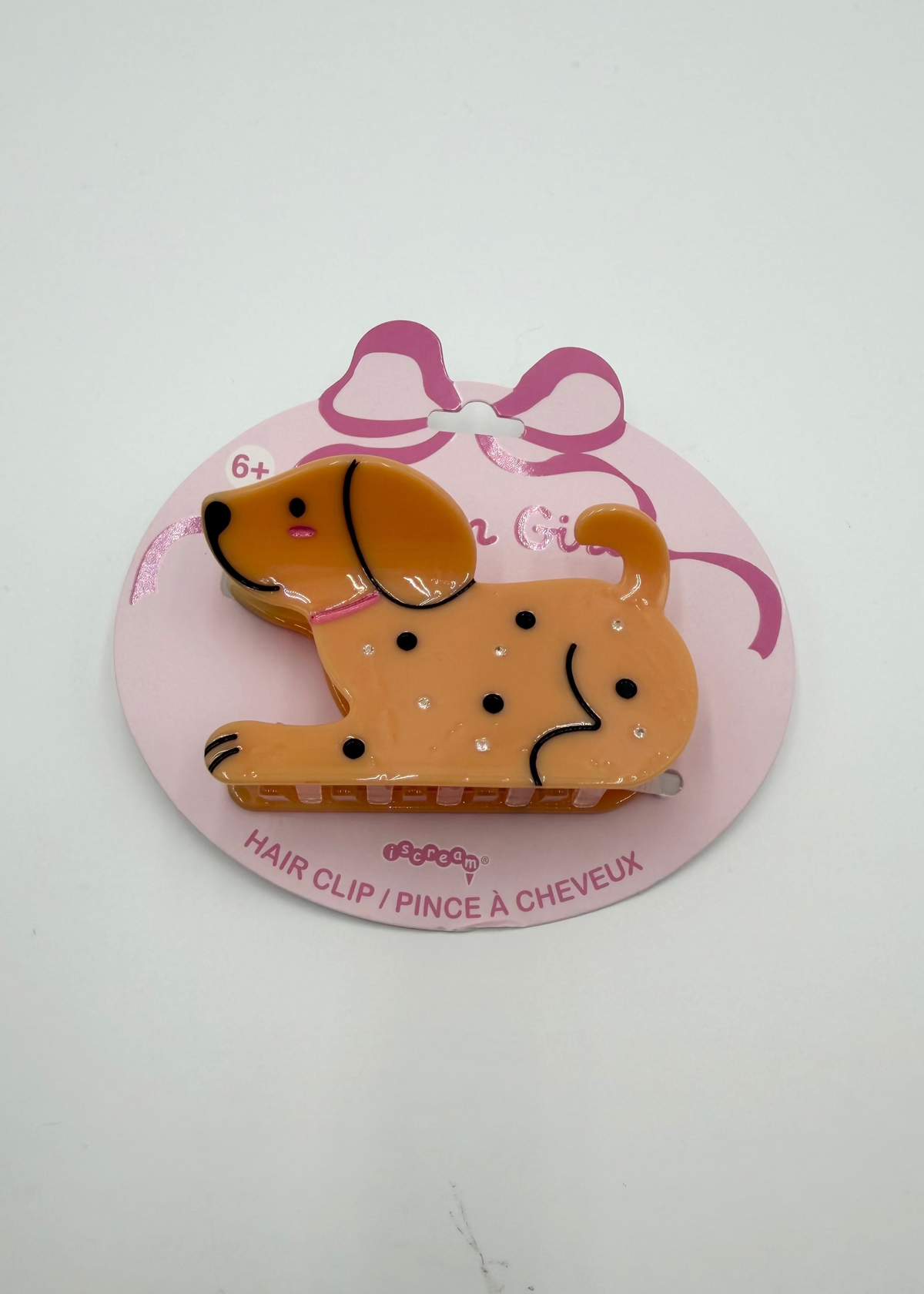 A dog-shaped hair clip in tan and brown with black details, displayed on a pink backing card with a bow illustration.