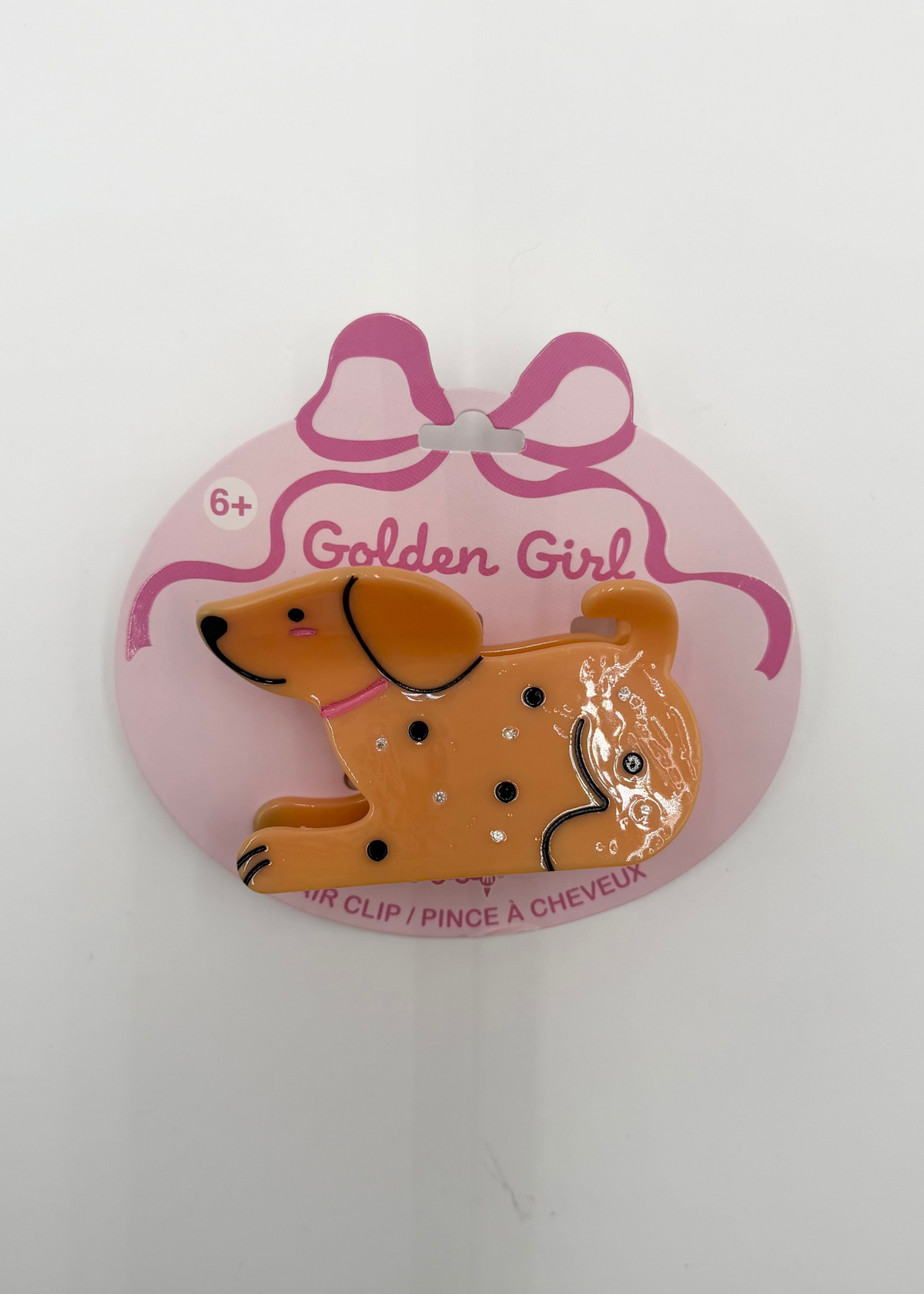 A Golden Girl dachshund hair clip attached to a pink oval cardboard backing with a bow illustration, photographed against a plain white background.