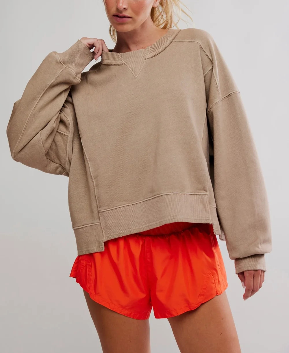 A woman wearing a beige oversized crewneck sweatshirt and bright orange athletic shorts, shown from the mouth down against a light gray studio background.