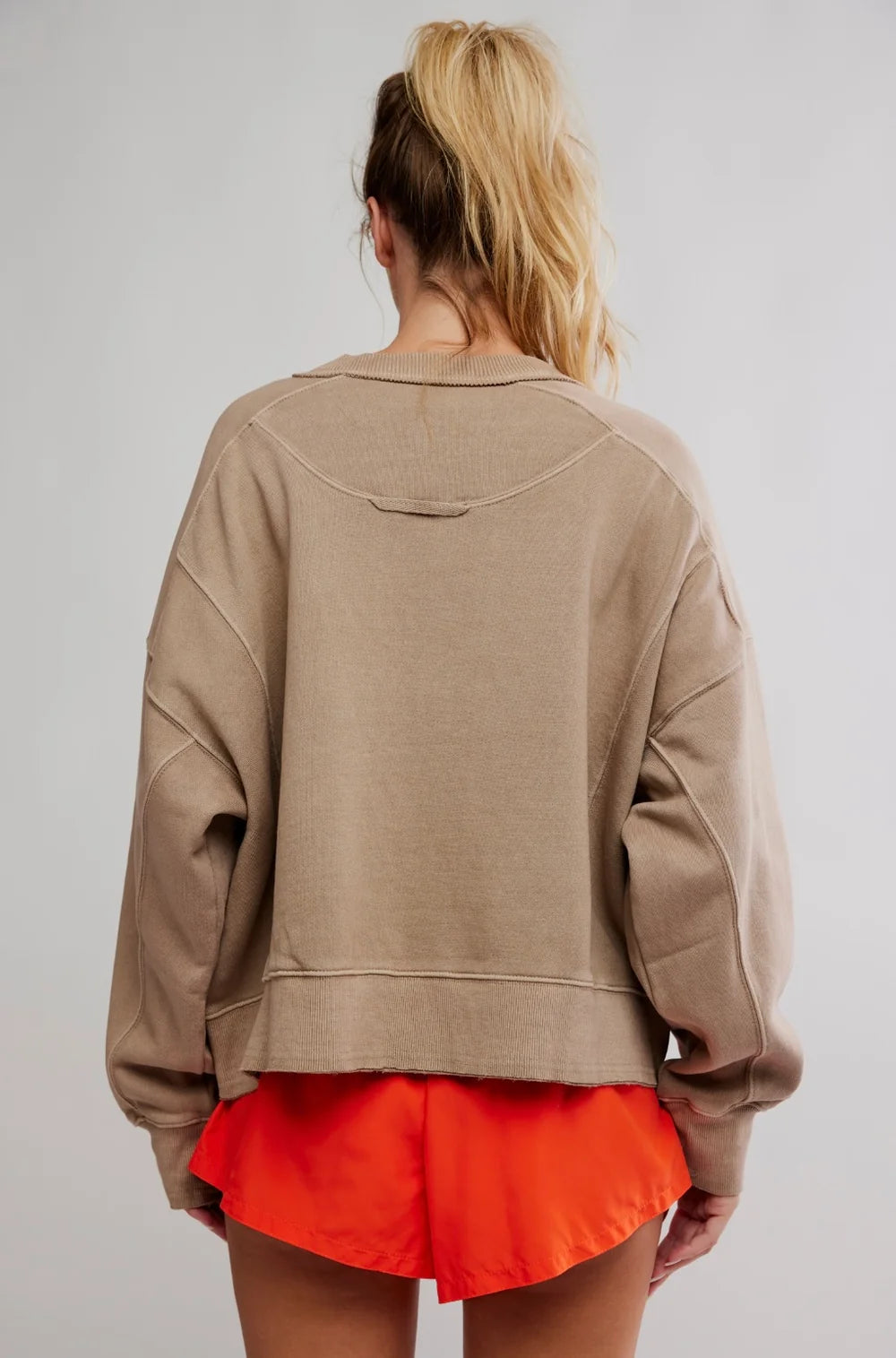 A woman shown from the back wearing an oversized taupe-beige cropped sweatshirt and bright orange shorts against a light gray studio background.