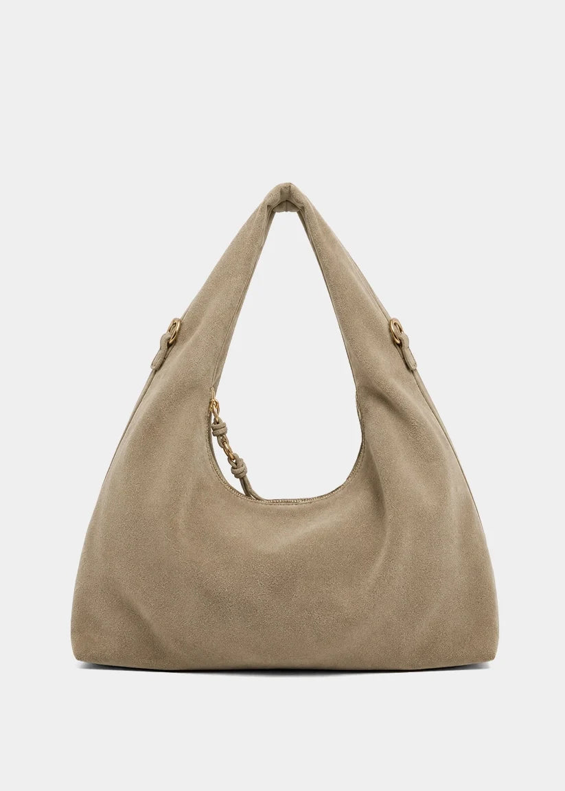 A slouchy hobo shoulder bag in taupe beige suede, featuring a knotted leather strap detail on one of the handles, set against a plain light gray background.