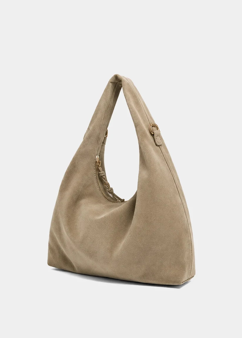 A slouchy hobo shoulder bag in taupe suede, featuring a single integrated shoulder strap and a small gold-tone ring detail on the side, set against a plain light gray background.