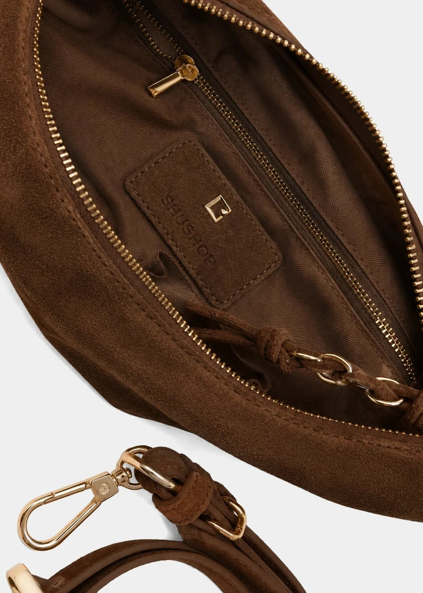 Close-up of a brown suede shoulder bag interior showing a zippered compartment, a leather brand patch, and gold-tone hardware on a light gray background.