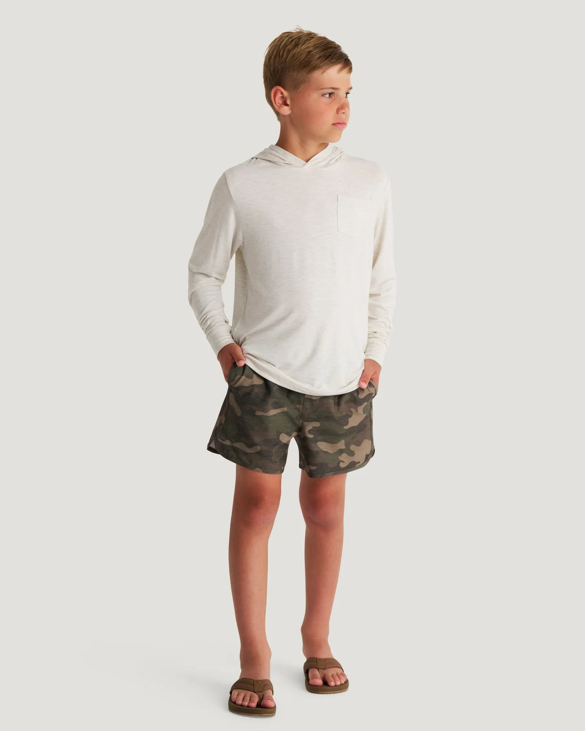 Young boy standing in front of a white background wearing a white and beige hoodie and camo shorts. 