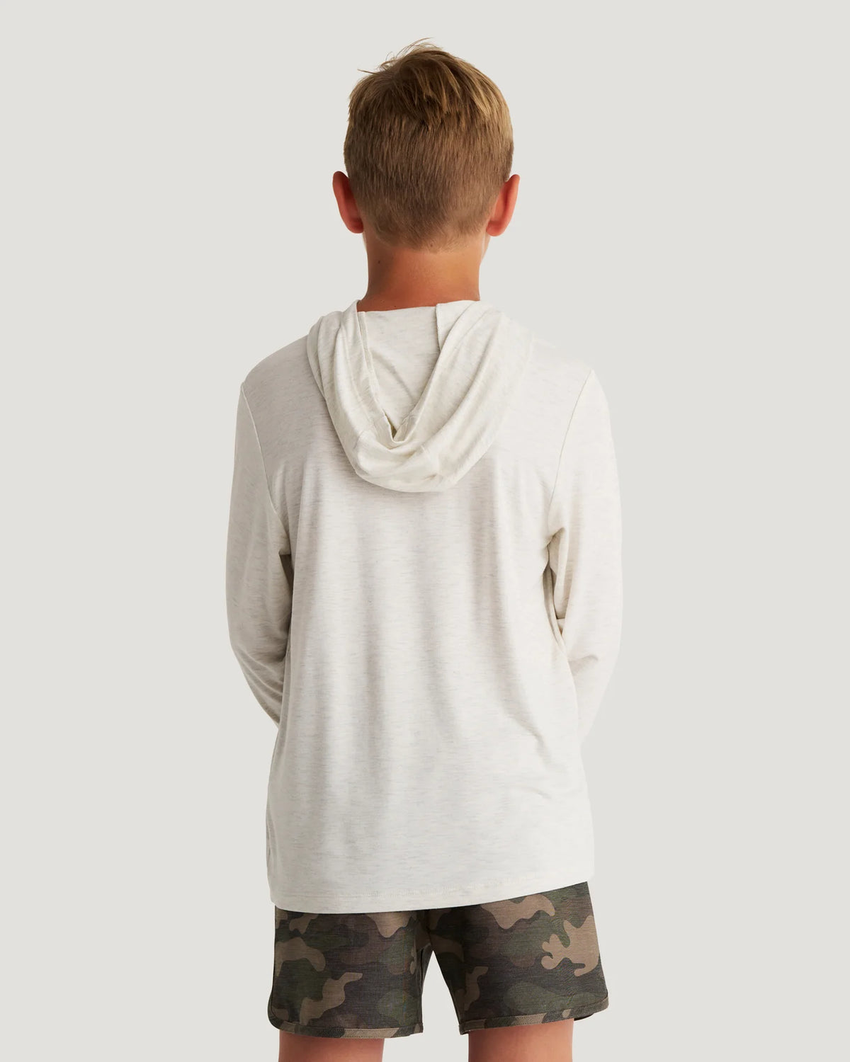 Back view of a young boy standing in front of a white background wearing a white and beige hoodie and camo shorts. 