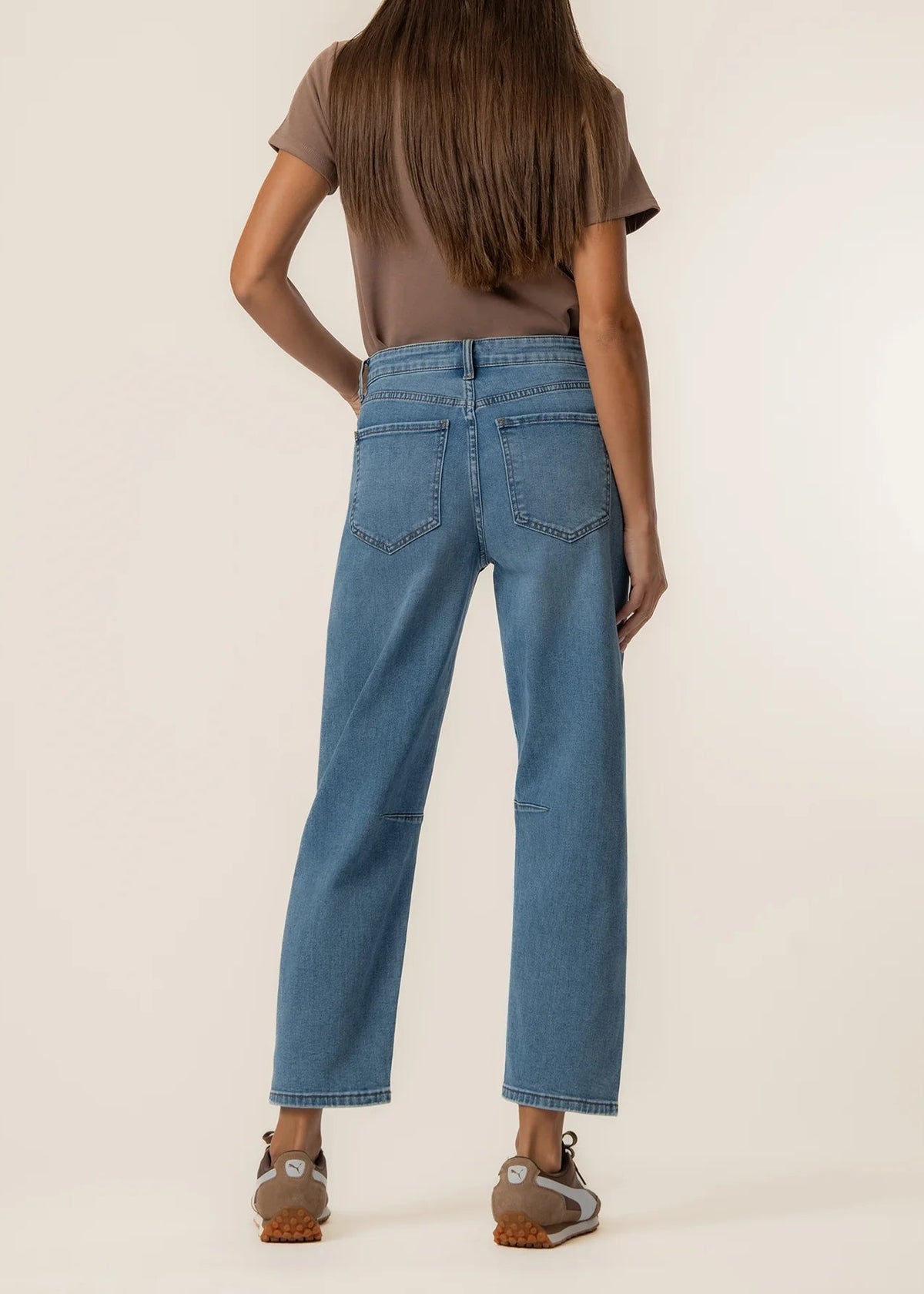 A model shown from the back wearing light blue cropped jeans, a brown t-shirt, and brown shoes against a plain beige studio background.