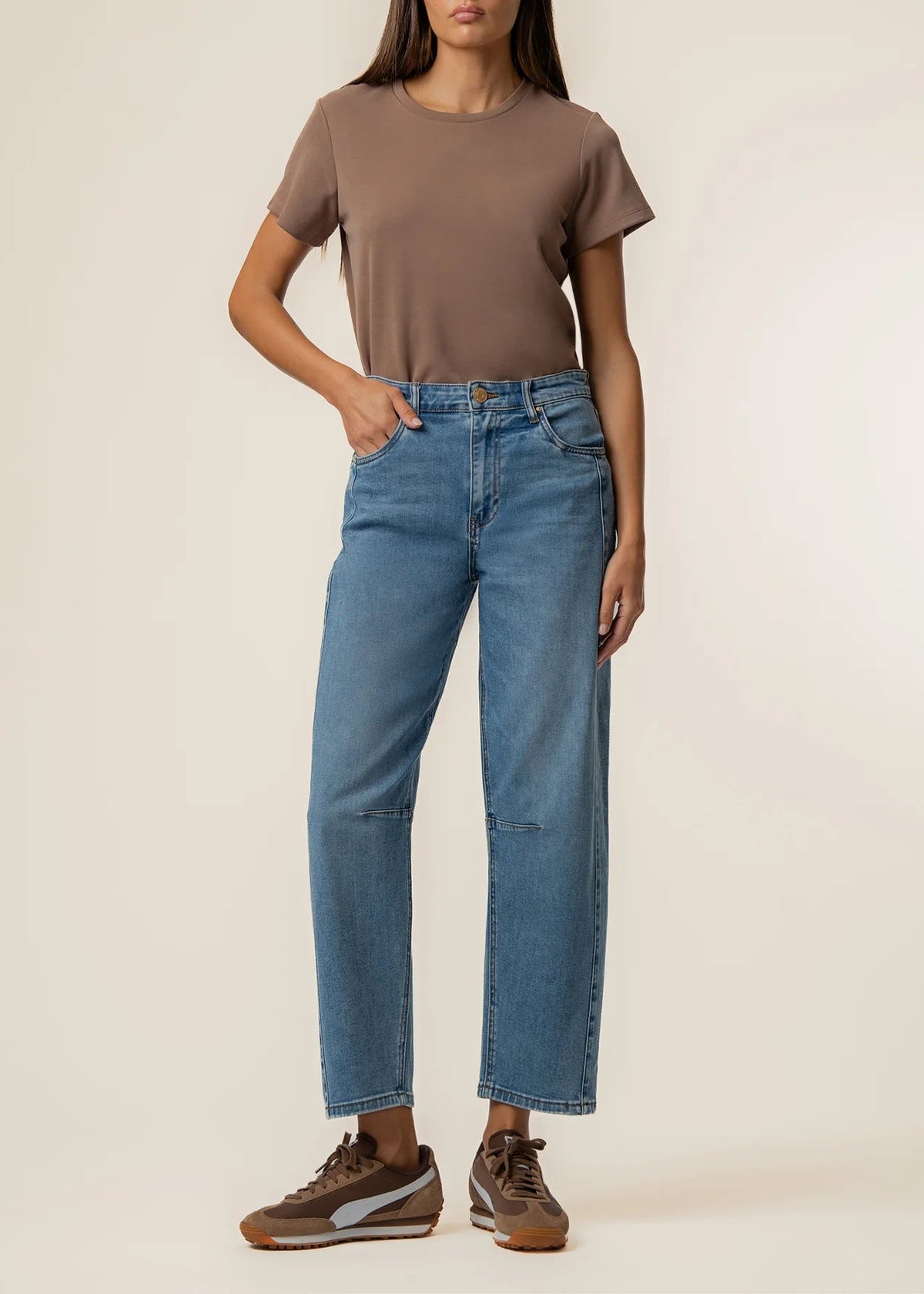 A model wearing a brown short-sleeve t-shirt, high-waisted straight-leg blue jeans, and brown sneakers, standing against a plain beige studio background.
