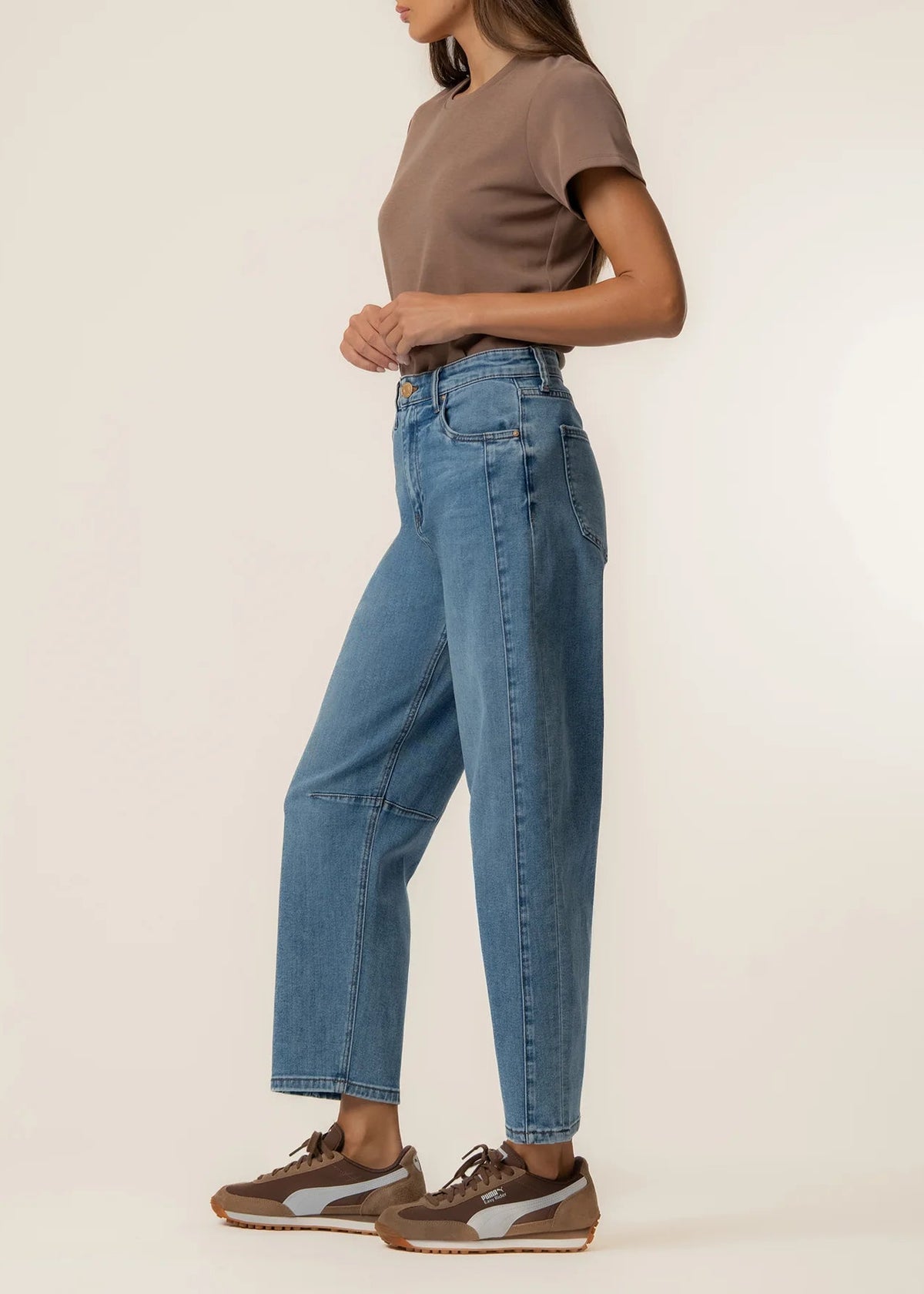 A side-profile view of a model wearing high-waisted, wide-leg blue denim jeans, a brown t-shirt, and brown sneakers against a plain off-white studio background.