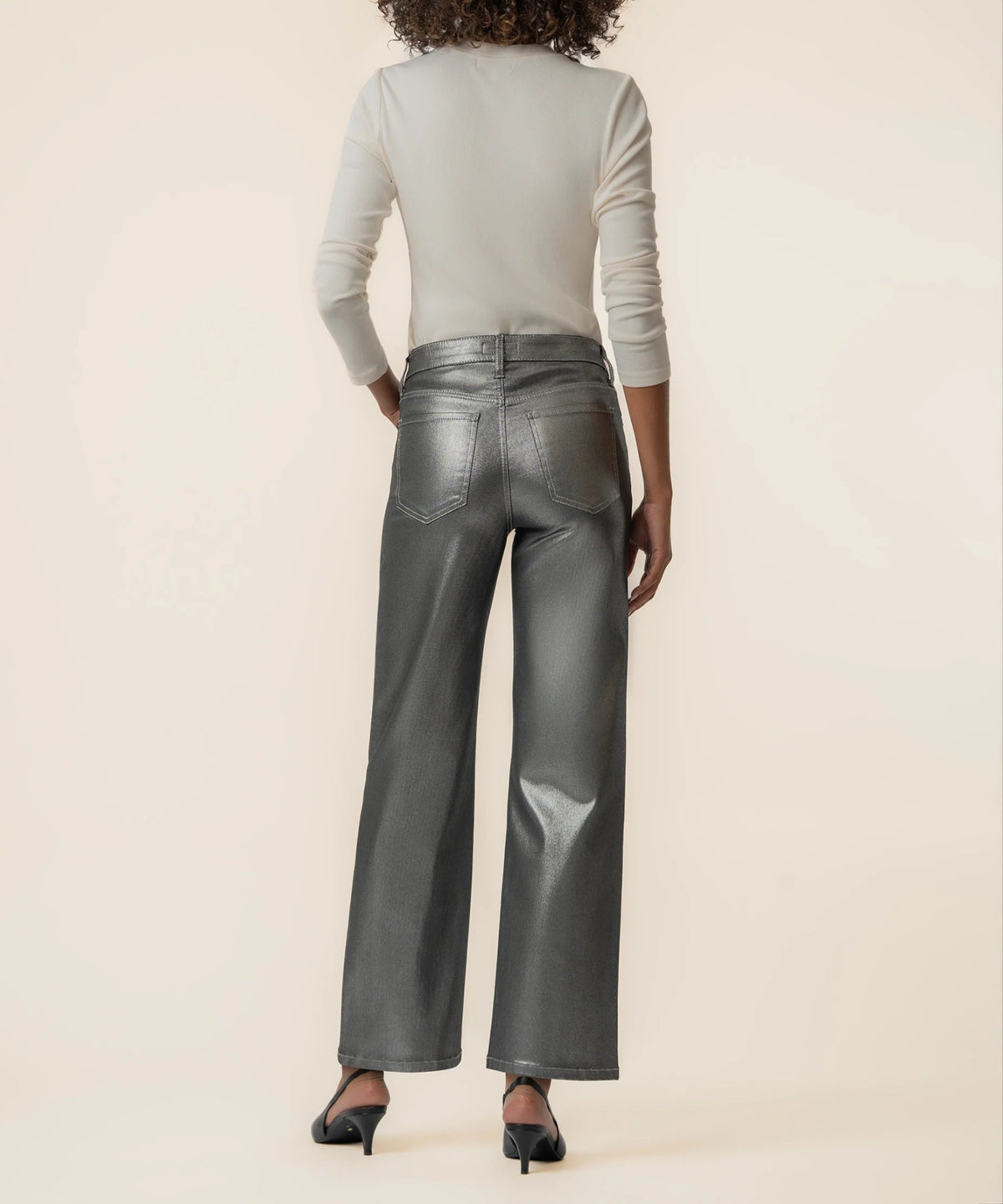 A model shown from the back wearing shiny dark gray wide-leg pants, a light beige long-sleeve top, and black heels against a solid beige background.