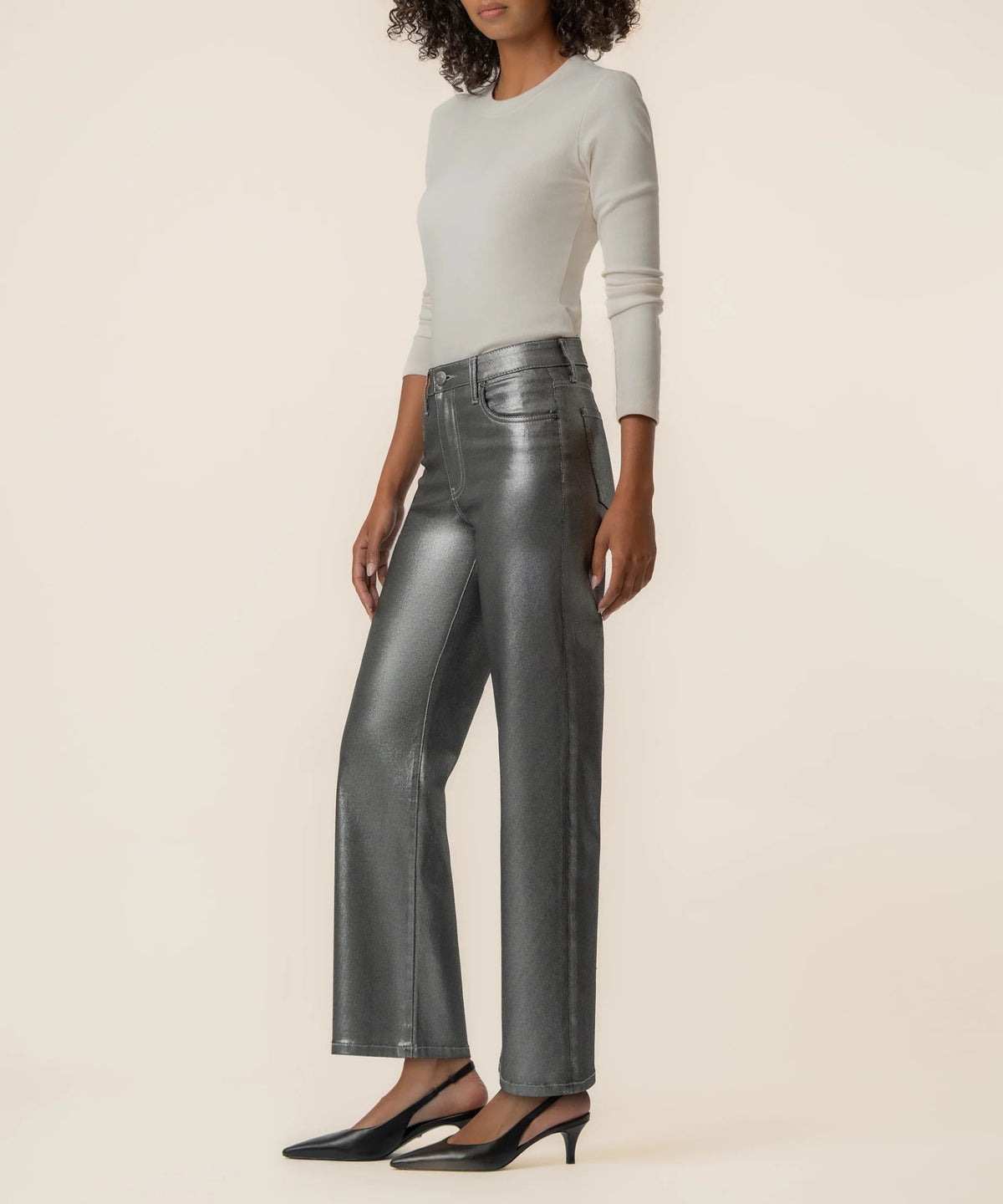 A model wearing high-waisted, wide-leg metallic gray pants, a light gray long-sleeve top, and black pointed-toe heels, standing against a solid cream background.