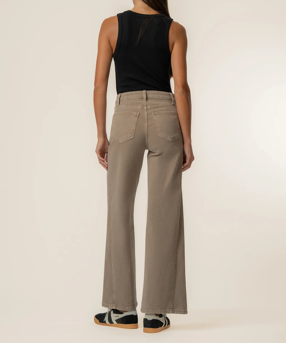 A model shown from the back wearing high-waisted, wide-leg taupe pants, a black sleeveless top, and black-and-white sneakers against a plain off-white studio background.