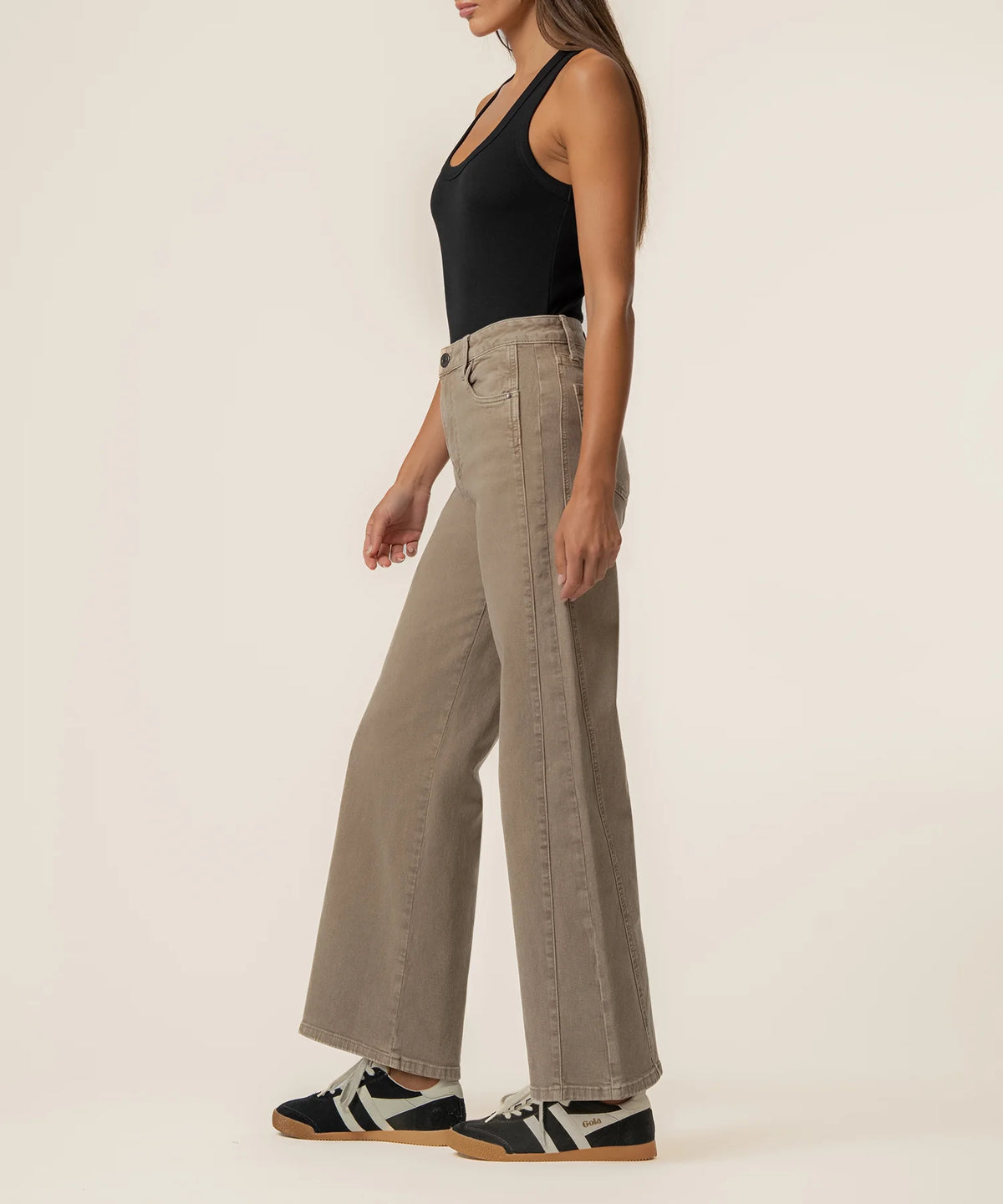 A woman shown from the side wearing high-waisted, wide-leg taupe pants, a black tank top, and black-and-white sneakers against a plain off-white studio background.