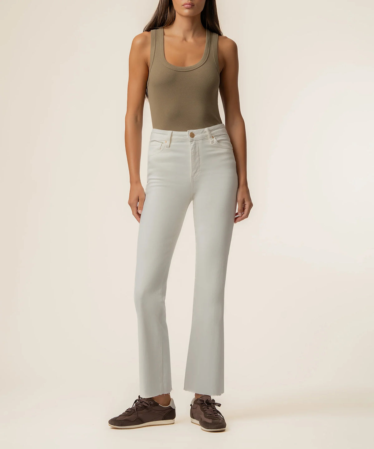 A model wearing light-wash, high-rise straight-leg jeans paired with an olive green tank top and brown suede sneakers, standing against a plain beige studio background.