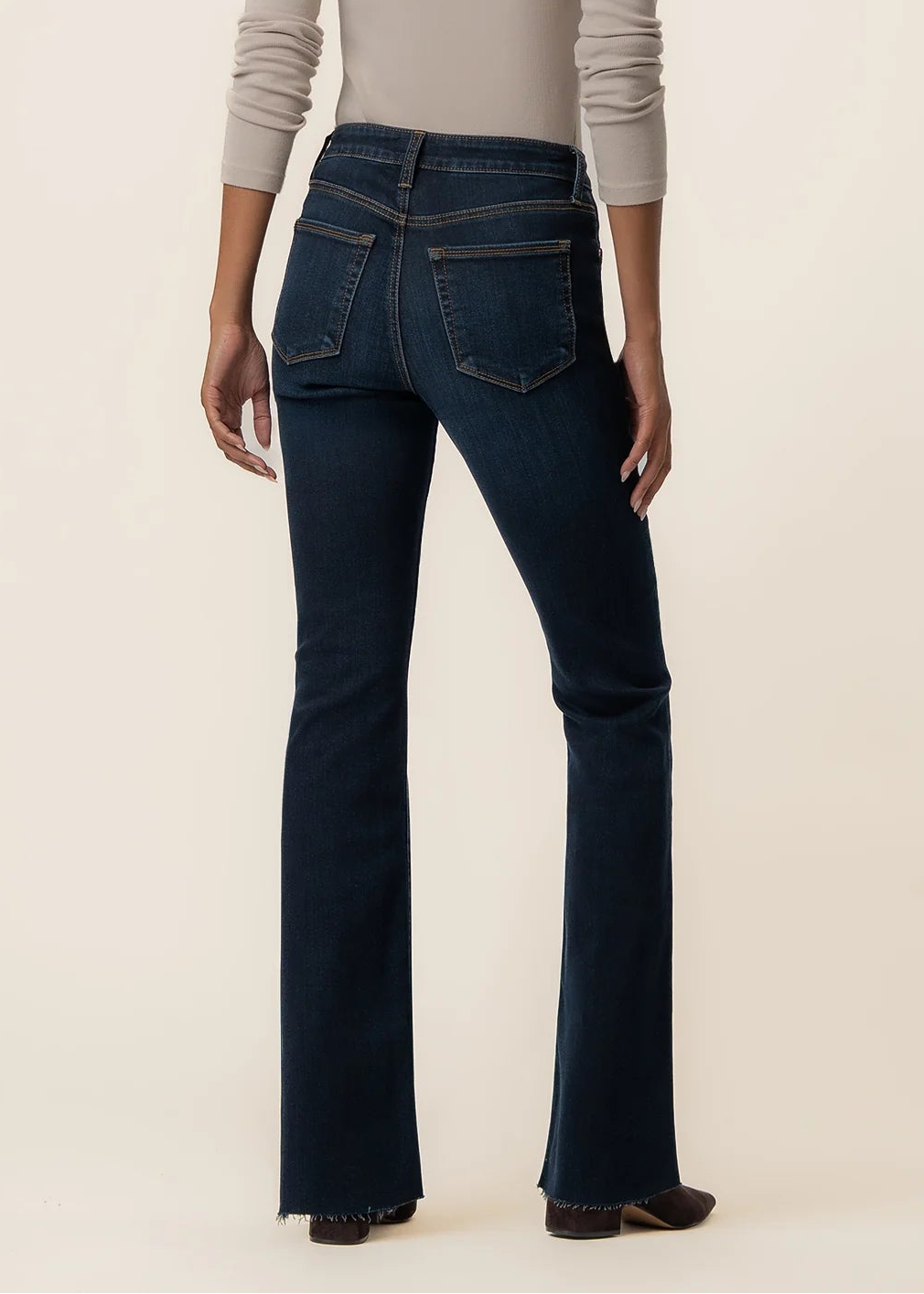 A model shown from the back wearing dark blue flared jeans and a light beige long-sleeve top, standing against a solid cream background.