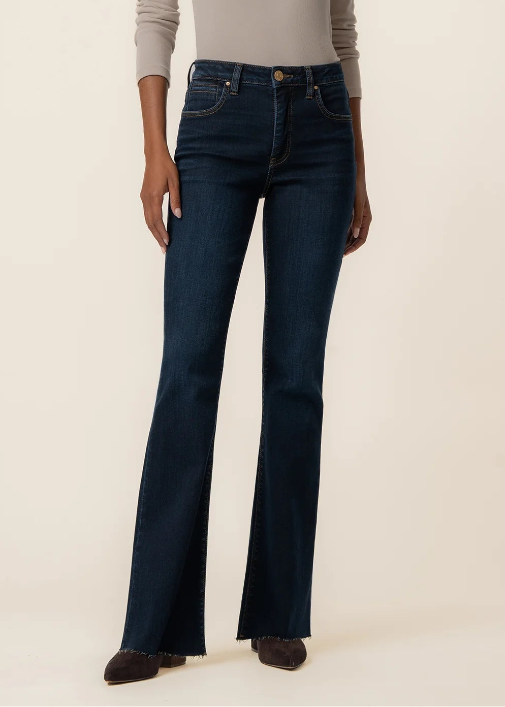 A model wearing dark blue high-waisted flare jeans with a beige long-sleeve top, standing against a plain beige studio background.