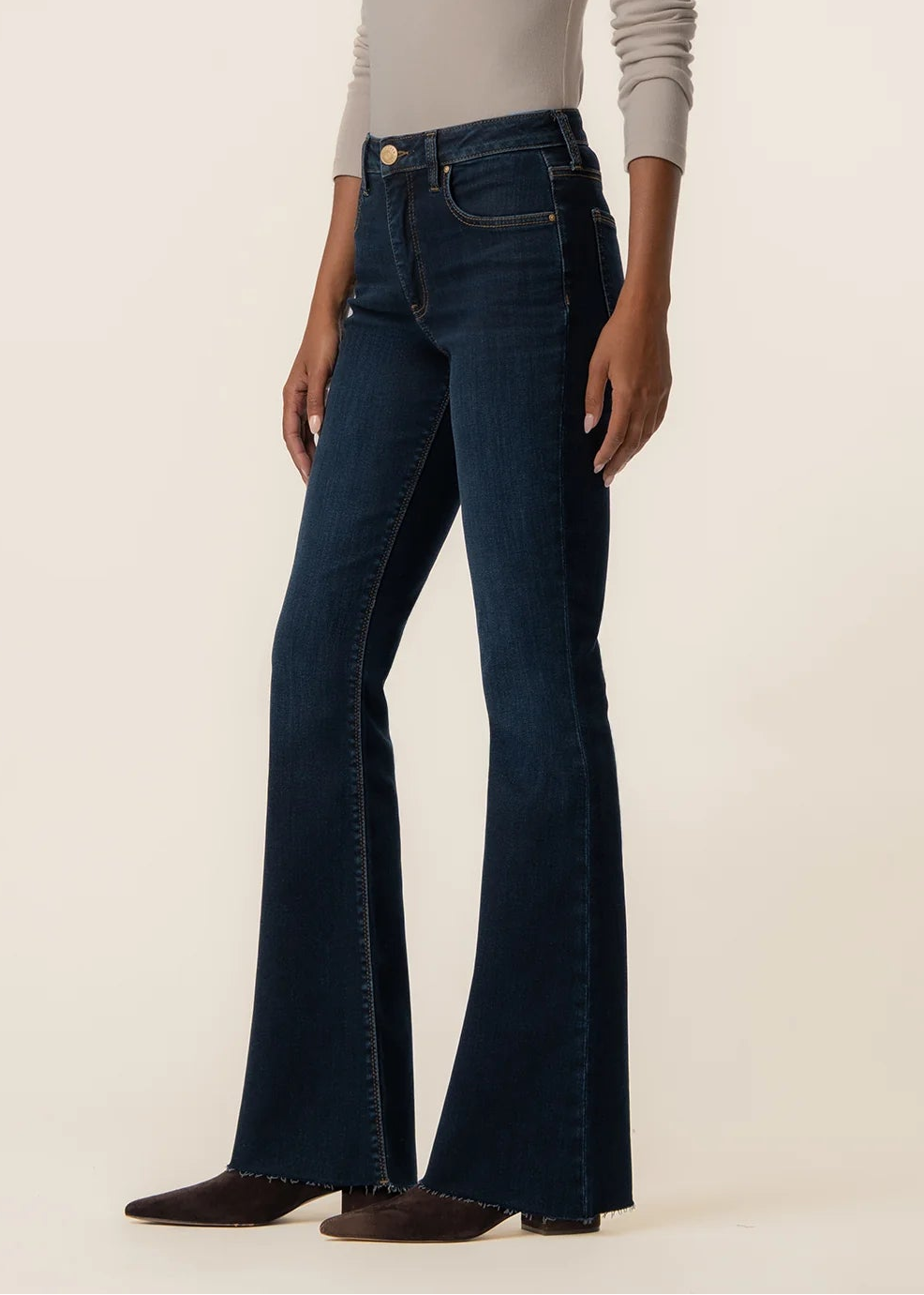 A model wearing dark blue high-waisted flare jeans with a beige long-sleeve top, standing against a plain beige studio background.